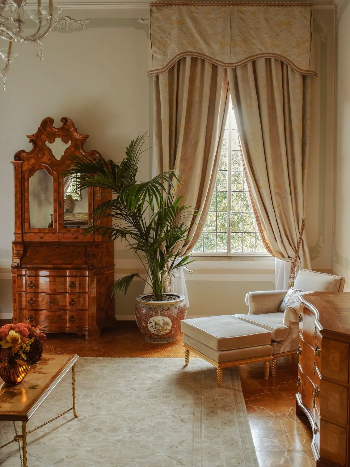 A vintage-style living room with a large window draped in ornate cream-colored curtains, a potted palm plant, an antique wooden dresser with a mirror, a beige upholstered chaise lounge, a coffee table with a floral centerpiece, and an elegant chandelier