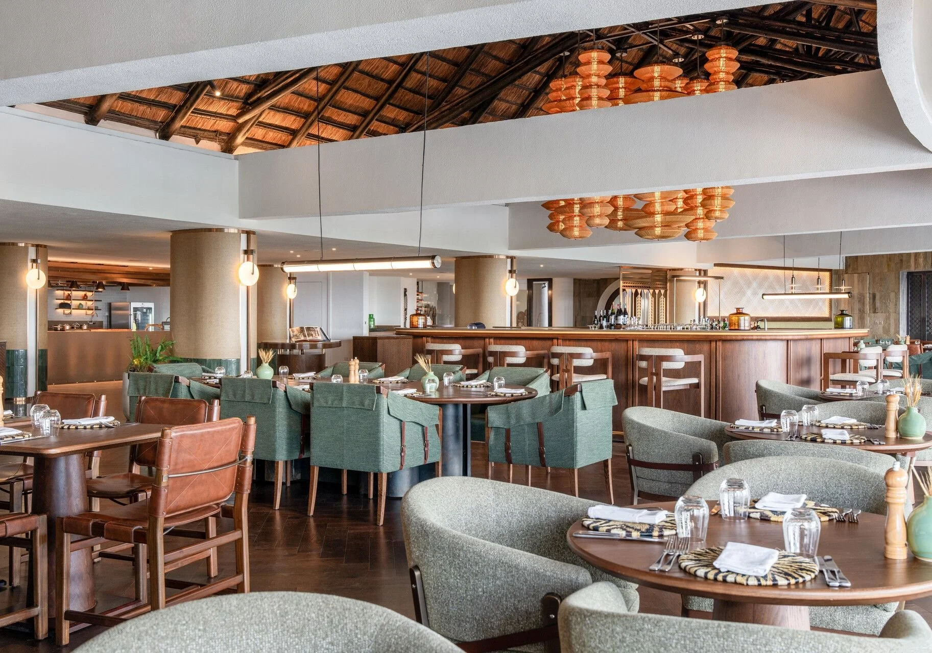 Upscale restaurant interior with wooden tables, green and brown upholstered chairs, and place settings with napkins and glassware. A wooden bar with barstools is in the background, with warm lighting and modern decor.