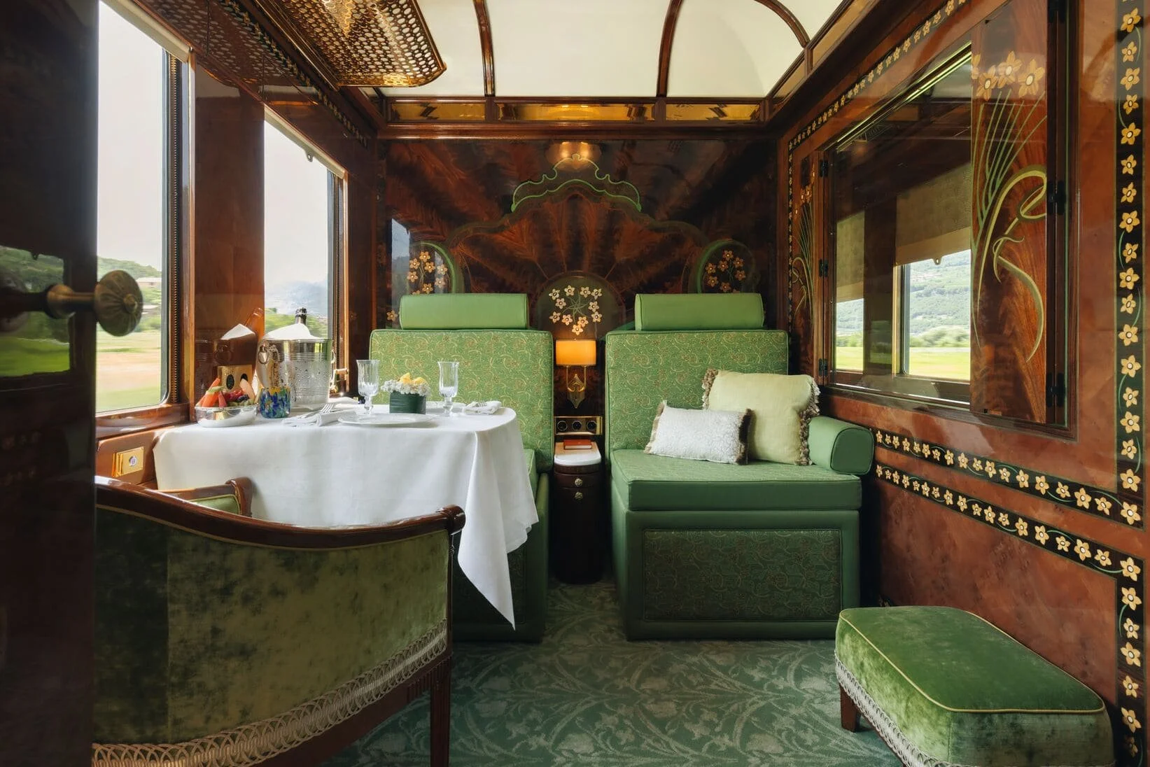 The interior of a vintage train dining car with ornate wood paneling, green upholstered seating, windows showing a scenic landscape, and a table set with white tablecloth, glassware, and breakfast items.