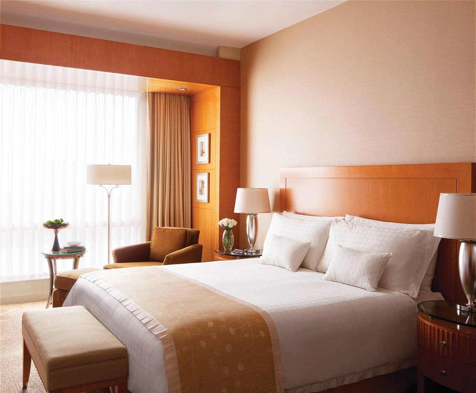 A hotel room with a large bed, nightstand with a lamp and a vase of flowers, armchair, side table with a fruit bowl, floor lamp, and window with sheer and drape curtains.