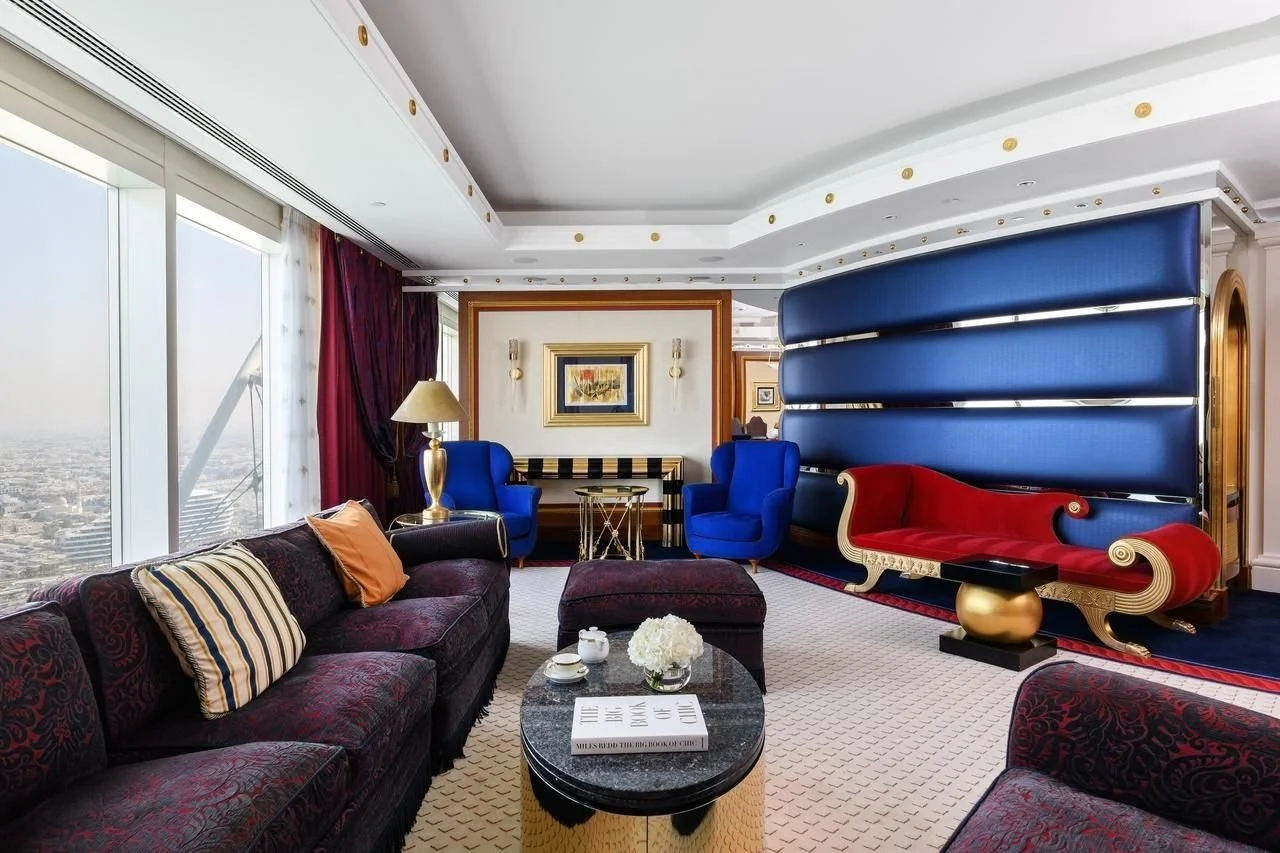 Luxurious hotel living room with dark purple sofas, gold and blue accents, large windows with city view, and stylish decor.