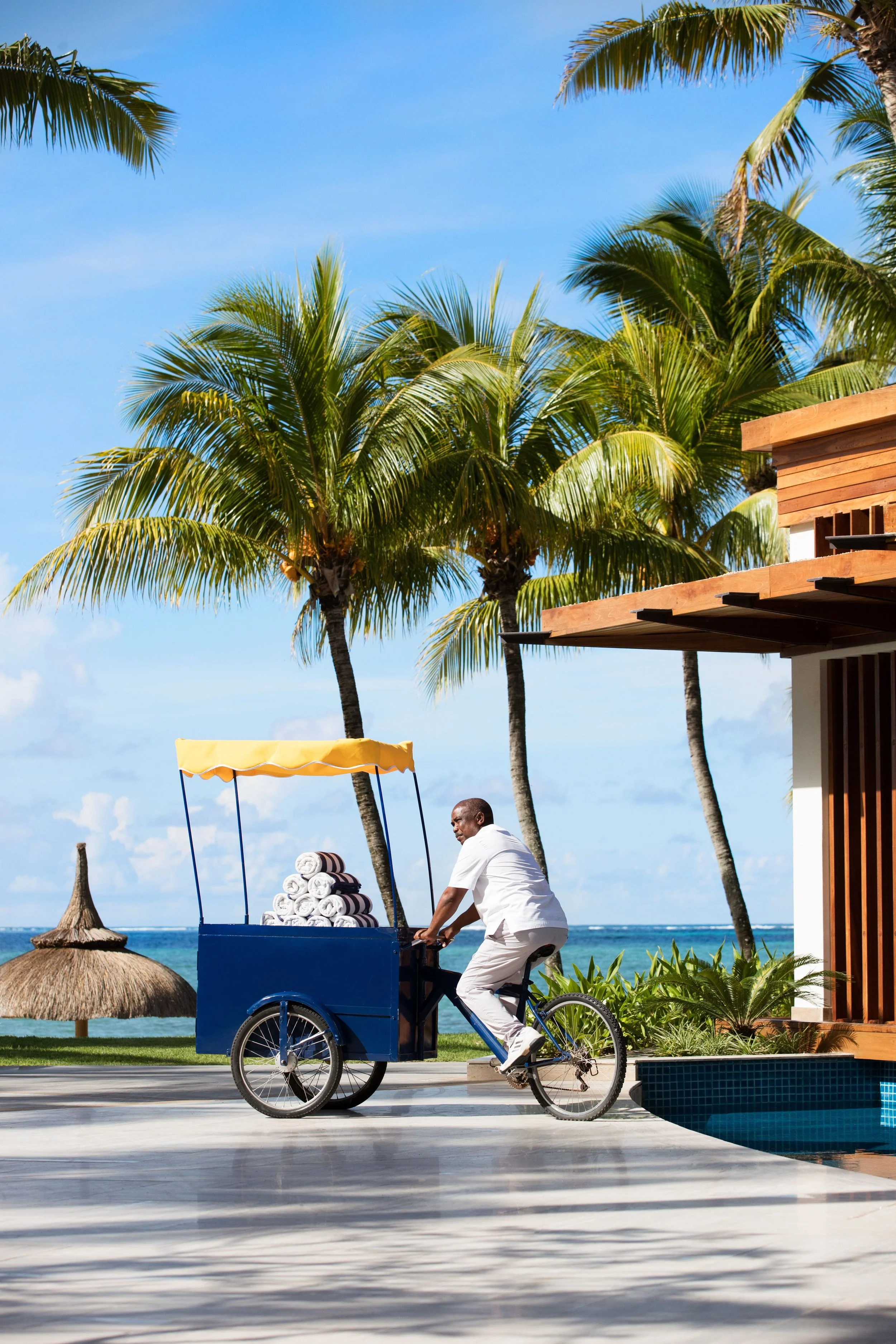 A man riding a blue bicycle with a side cart filled with rolled towels along a beachside promenade, with palm trees, thatched umbrellas, and a blue sky in the background.