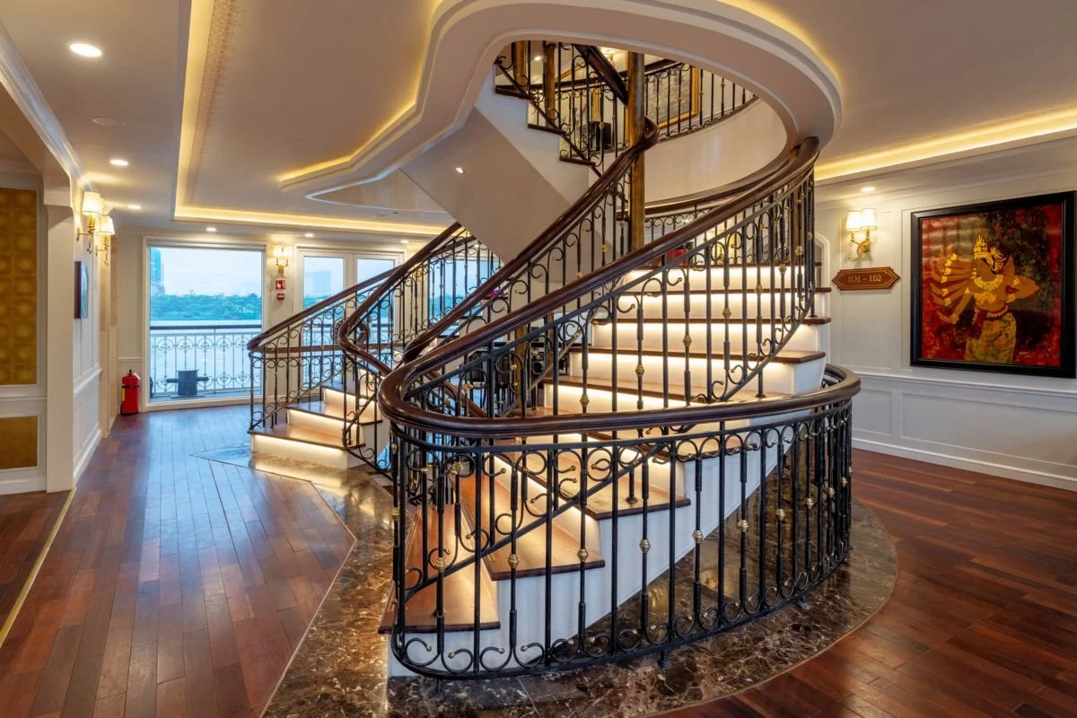 Interior view of a staircase with a wooden handrail and ornate black metal railing, illuminated with soft lighting, in a luxurious setting with hardwood floors and framed art on the wall.