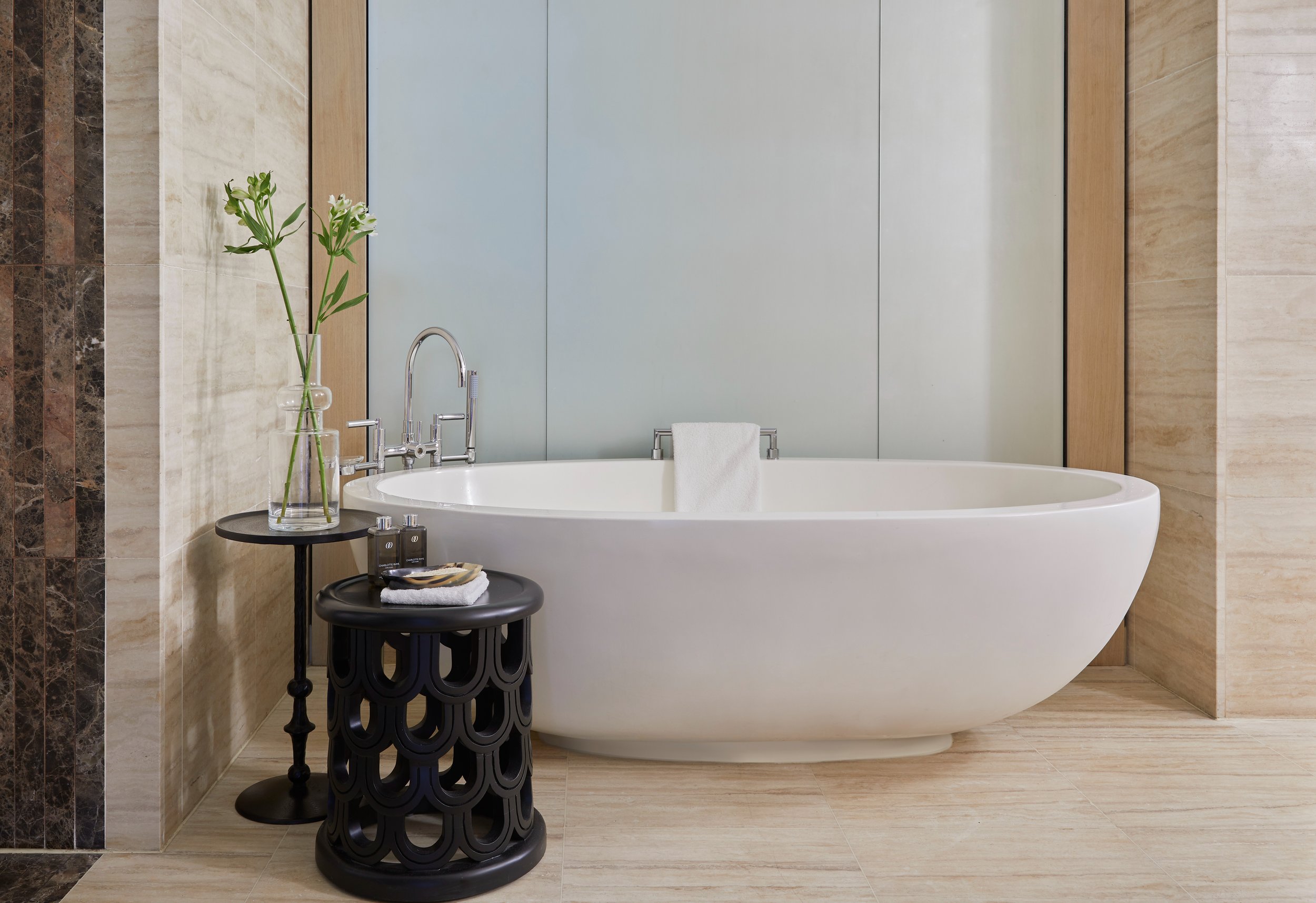 A bathroom with a modern, freestanding white bathtub positioned against a wall with light beige tiles. To the left, a black round side table holds a glass vase with white flowers, bath products, and a rolled white towel. The wall behind the tub features a large vertical gray panel with wooden trim on either side.