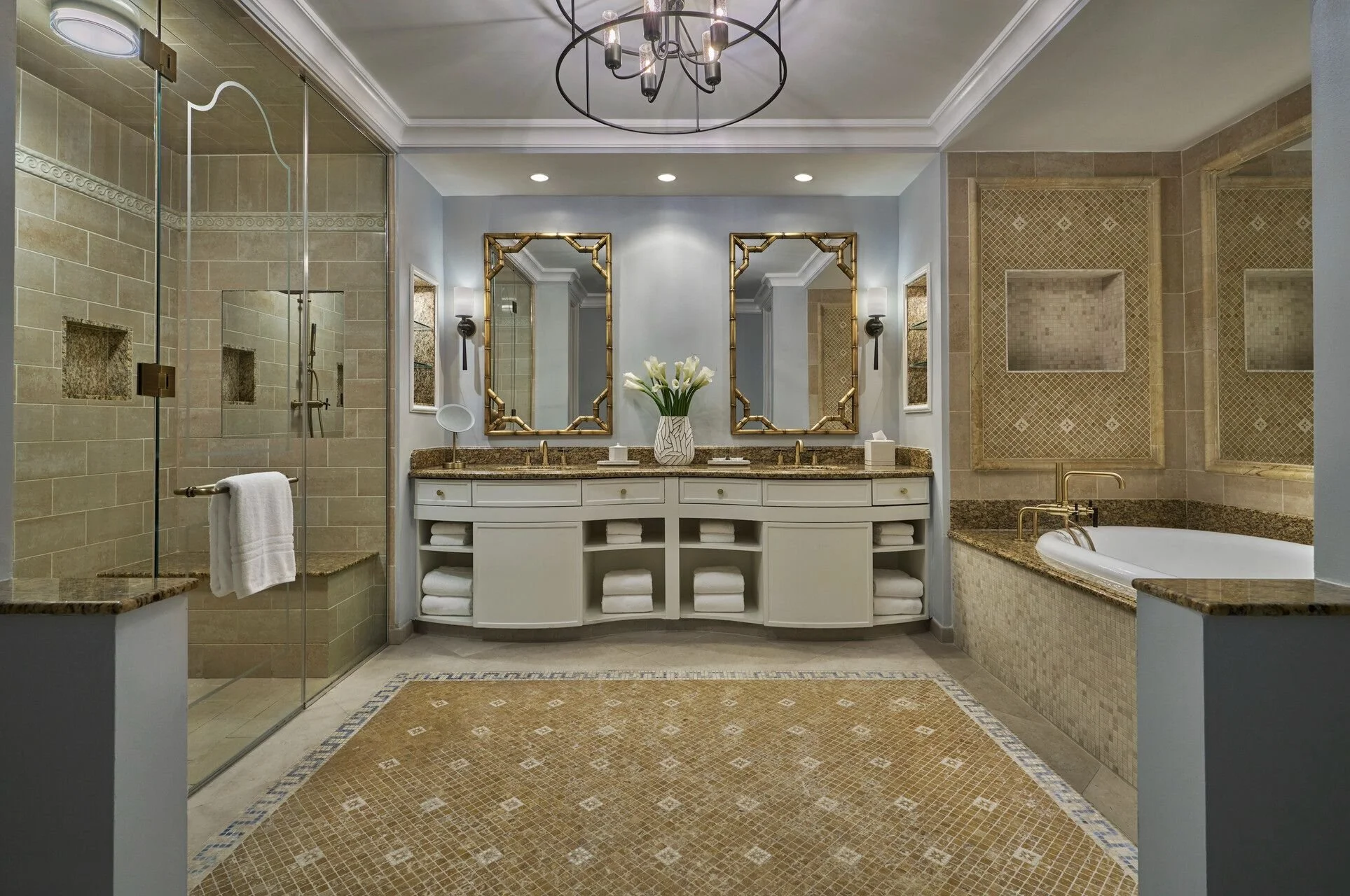A luxurious bathroom featuring a double vanity with two mirrors, a large soaking tub, a shower enclosed with glass doors, and elegant beige tilework, accented by a modern chandelier and decorative lighting.