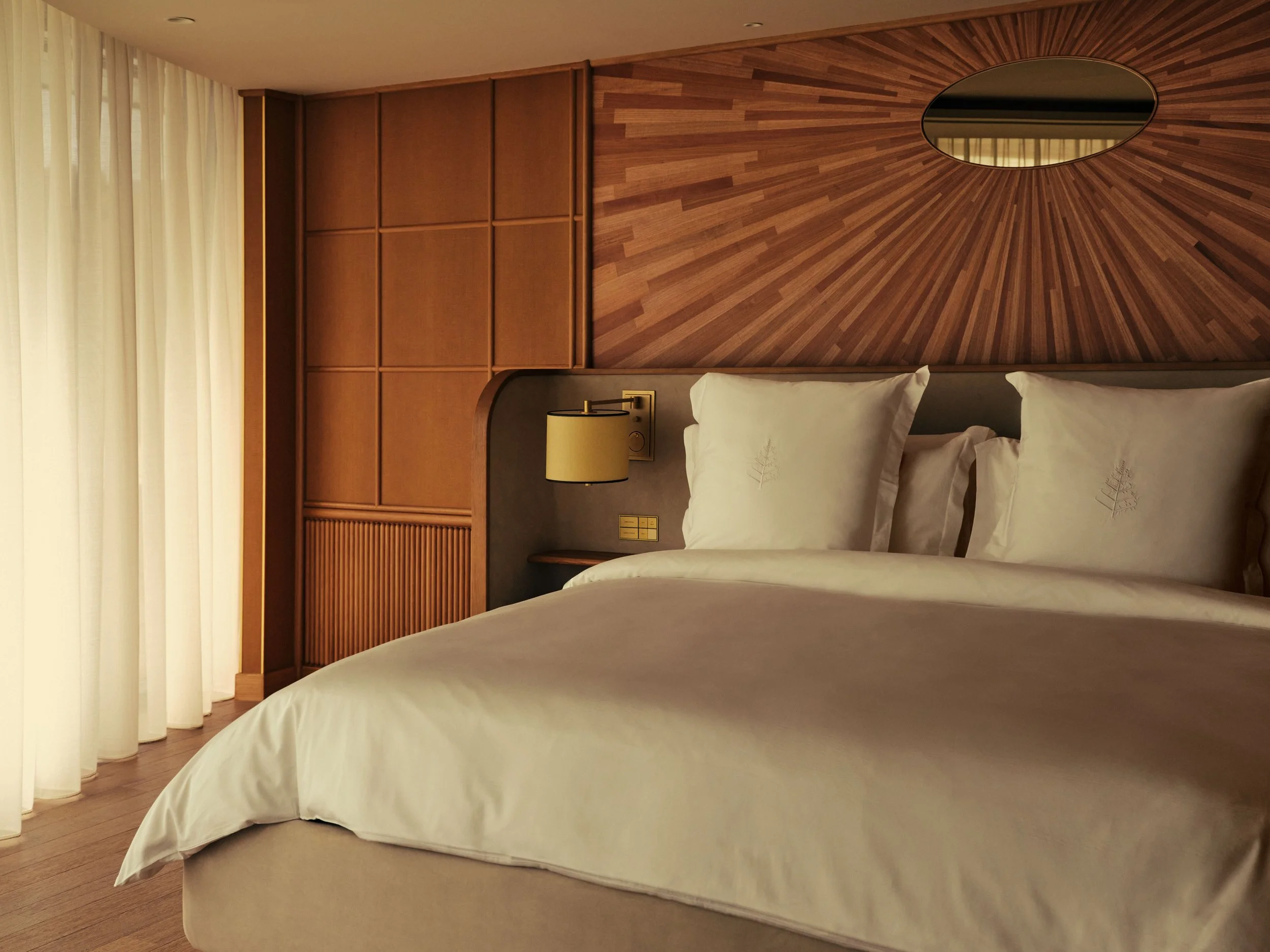 A bedroom with a large bed, white bedding, and a gray headboard. There is a nightstand with a lamp next to the bed. The wall behind the bed features a wooden panel with an oval mirror.