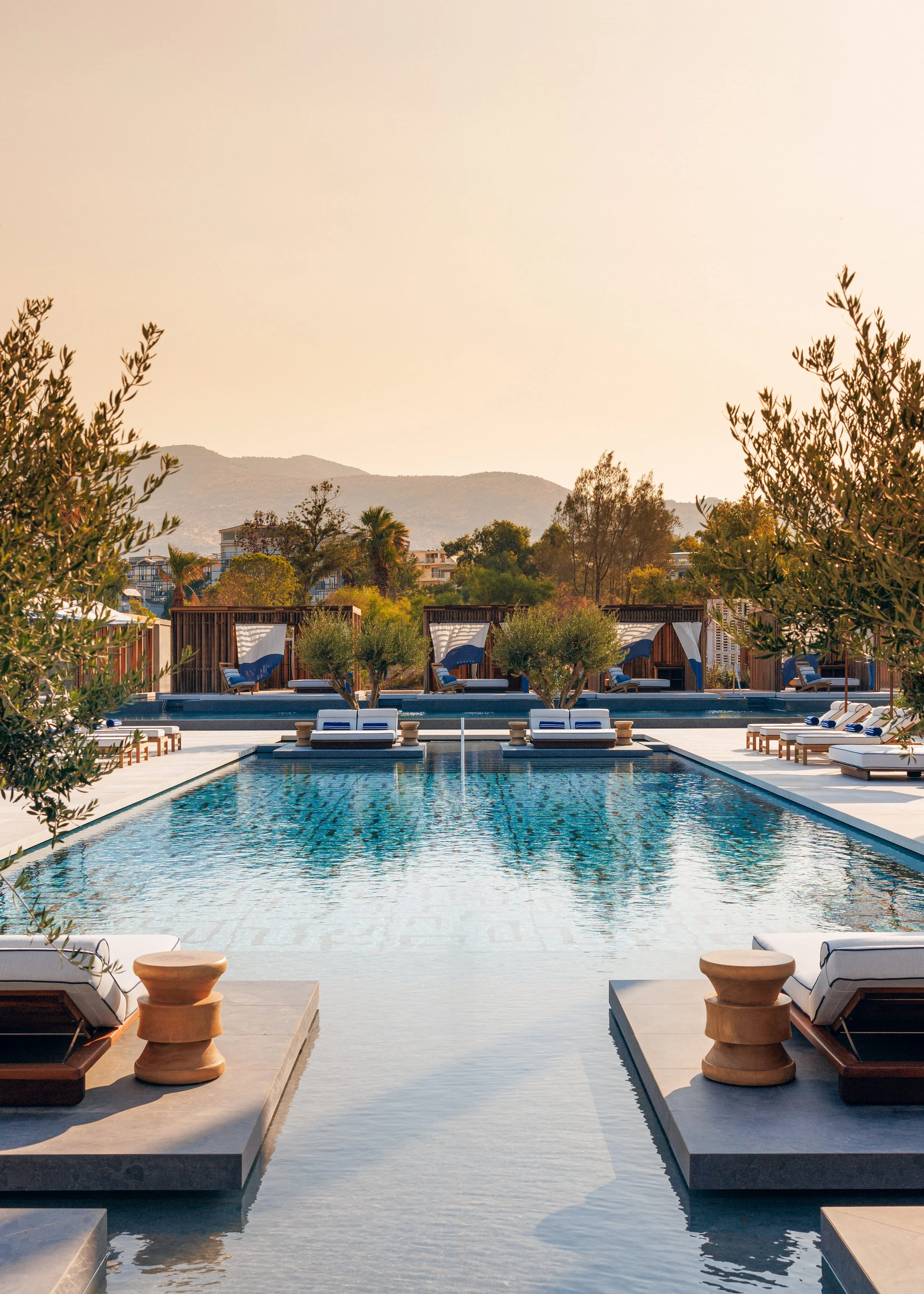 A luxurious outdoor swimming pool area with lounge chairs, cabanas, and tall trees, with mountains in the background during sunset.