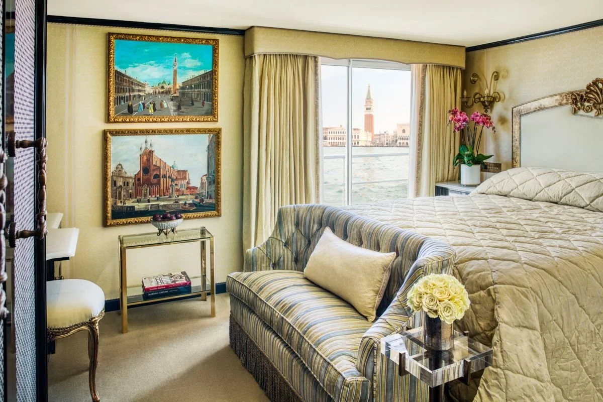Luxury hotel room with a large bed, striped armchair with pillow, glass side table with white roses, paintings on yellow wall, and views of Venice with water and historic buildings.