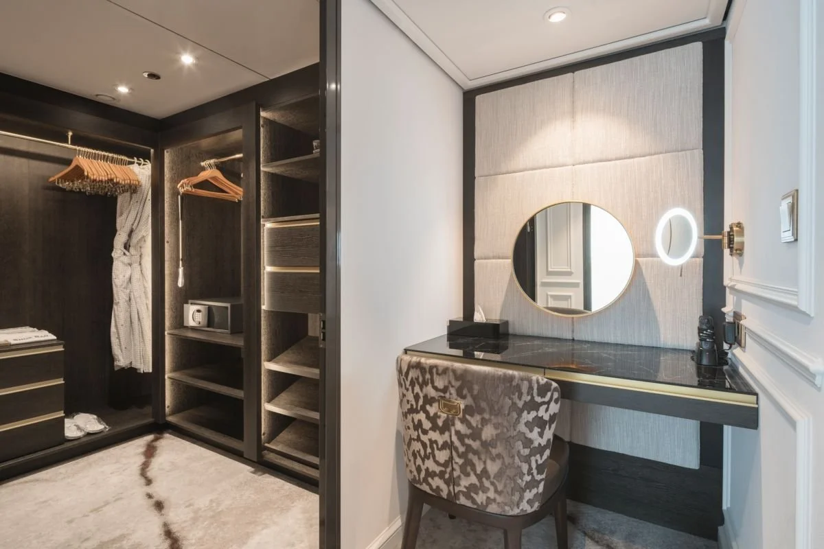 Closet with dark wood finish and open shelves, some hanging clothes, a safe, and a drawer on the left. A makeup or work vanity with a round mirror, a lighted magnifying mirror, and makeup or grooming items on the right. The space is modern and luxurious.
