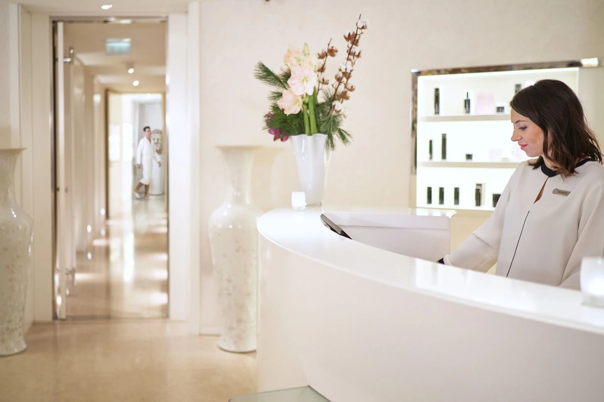 Hotel reception with a woman staff member sitting at the desk, white walls, a tall flower arrangement, and a hallway in the background with a person walking.
