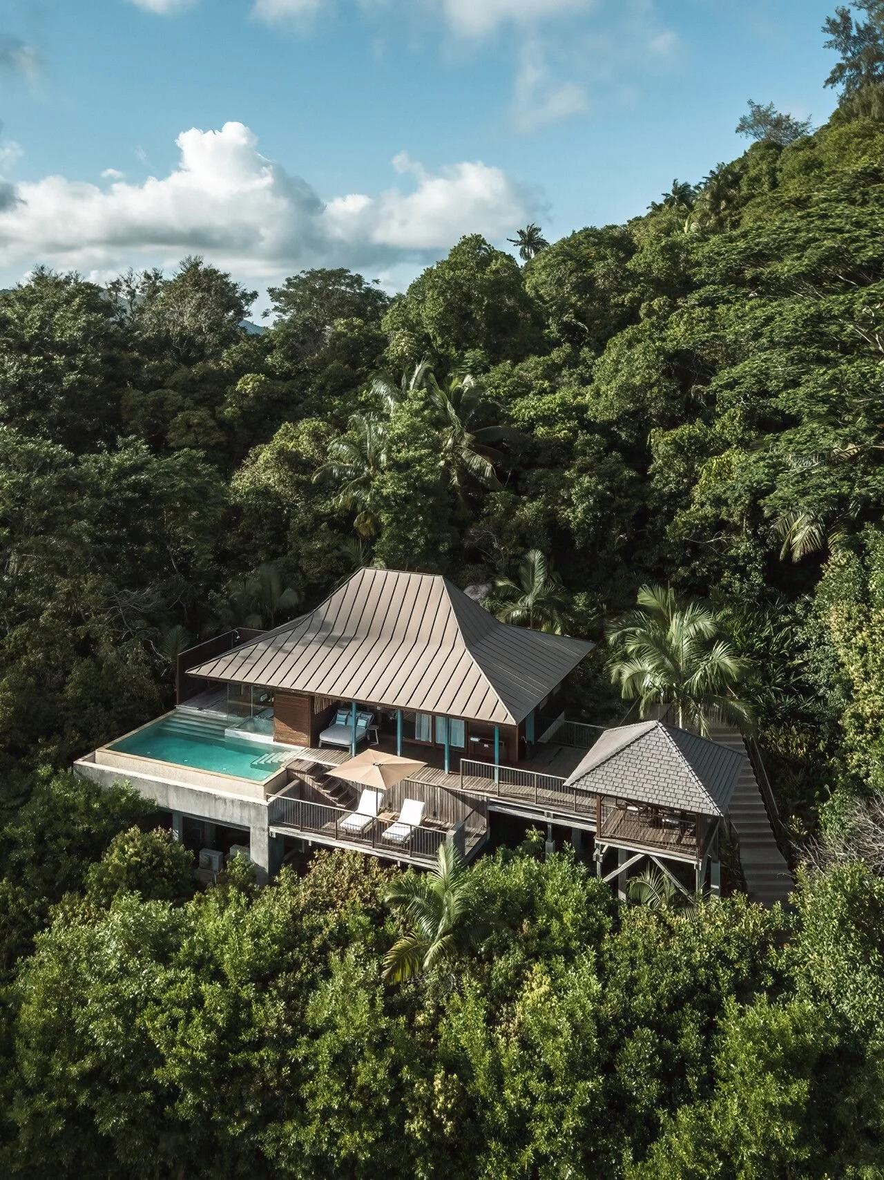Luxury house with swimming pool surrounded by lush tropical forest under blue sky with clouds.