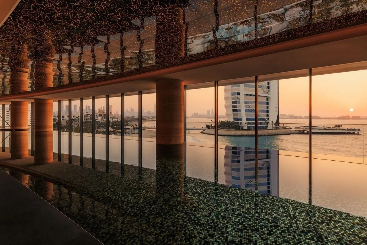 Modern indoor space with large glass windows overlooking a city skyline and water at sunset, with a reflective ceiling, wooden columns, and a mosaic-tiled floor.