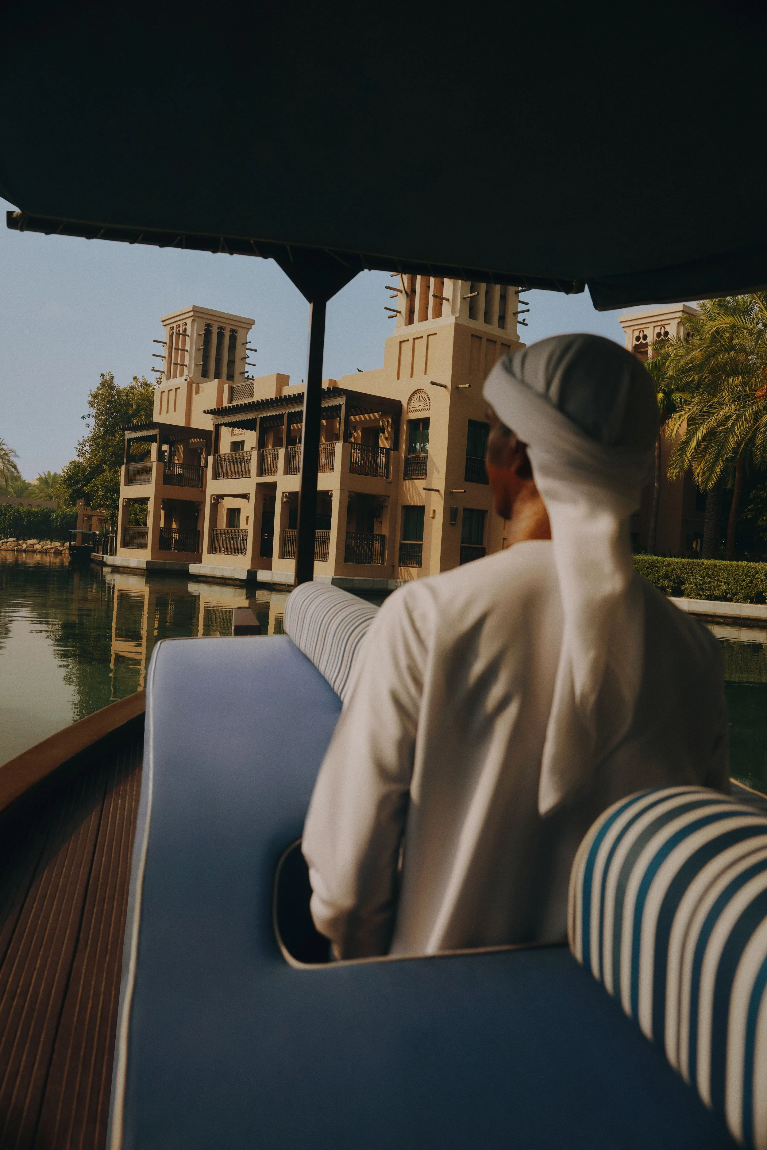 A person wearing a white traditional Middle Eastern outfit and head covering is sitting on a boat. The boat is on a canal with a beige building with multiple balconies and vents in the background. There are some palm trees and water reflection visible.