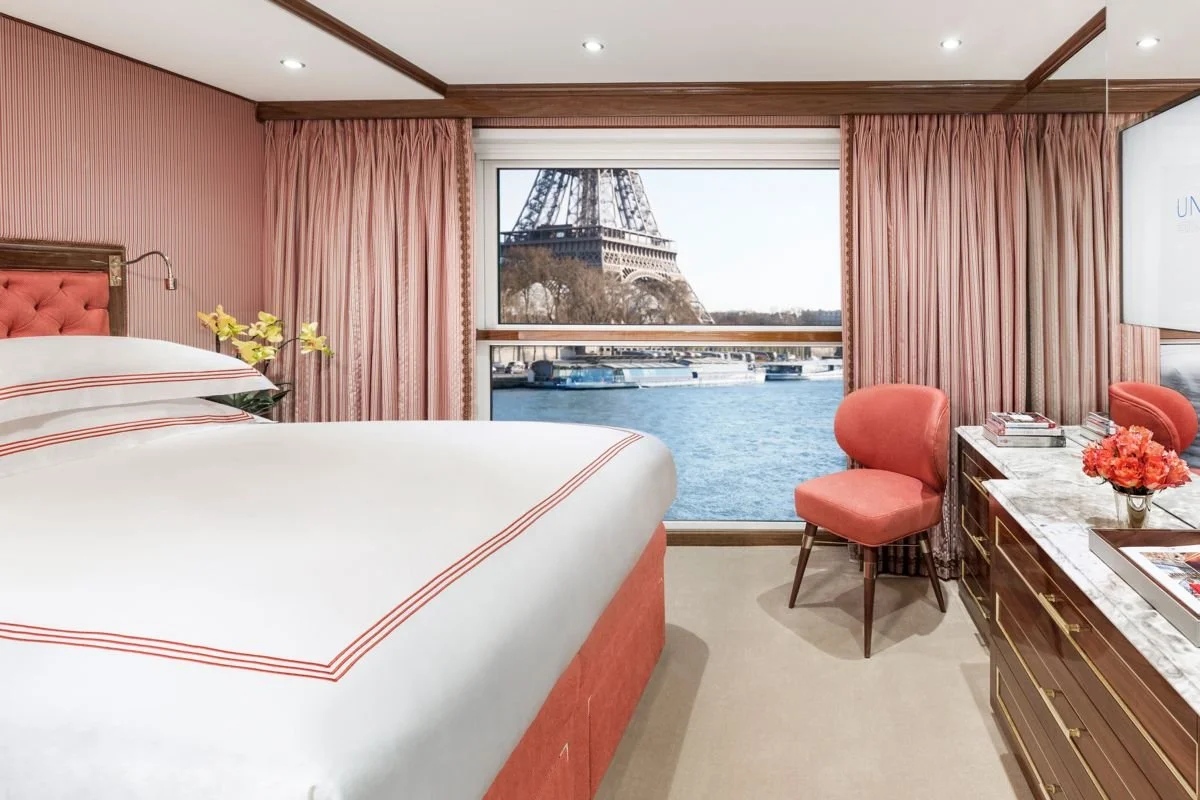 A hotel room with pink curtains, a large window showing the Eiffel Tower and the Seine River, a bed with white and pink striped bedding, pink chairs, and a marble-topped dresser with flowers and books.