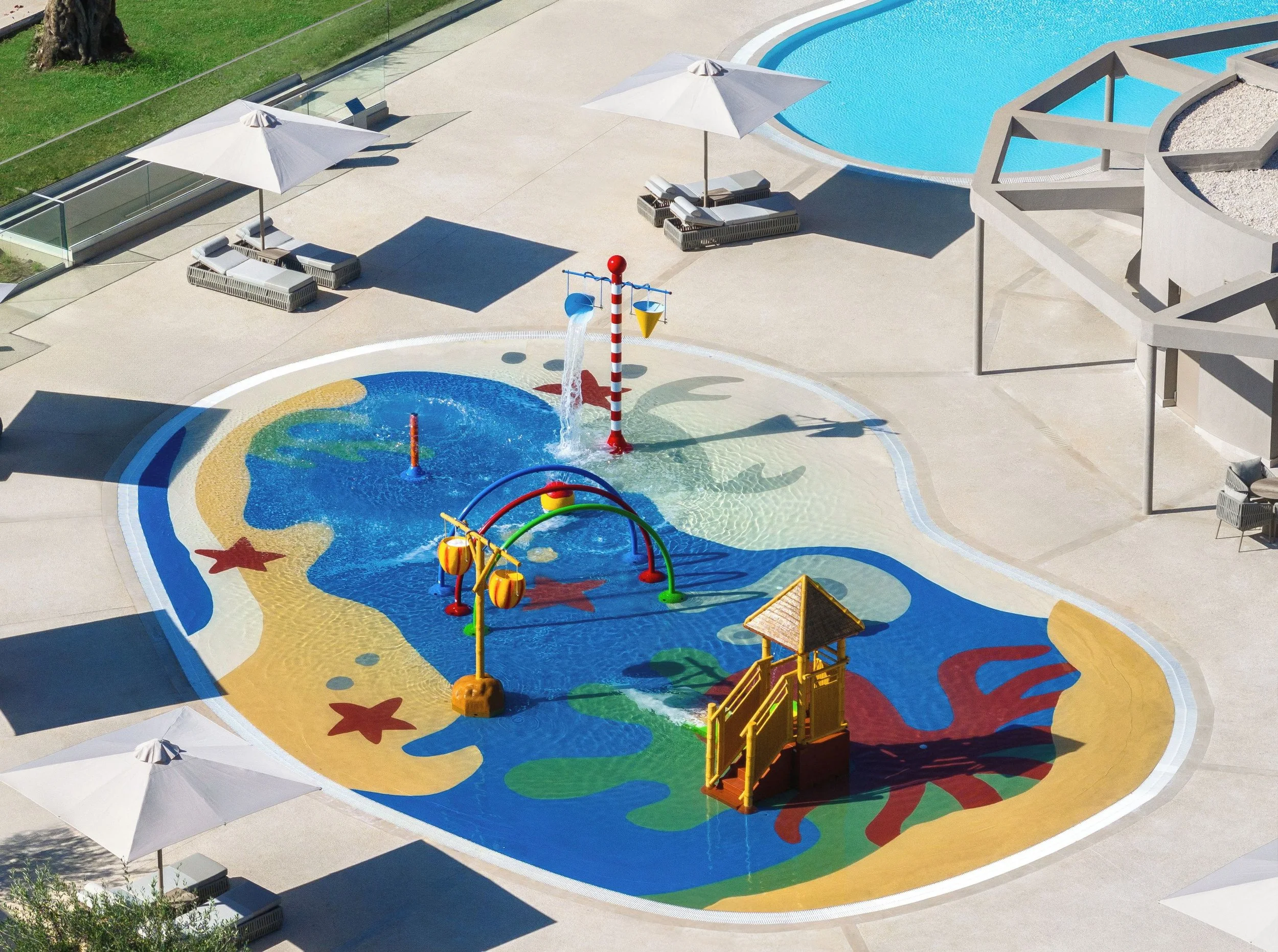 A colorful splash pad with water features, surrounded by lounge chairs with umbrellas, a pool, and a grassy area with a tree.