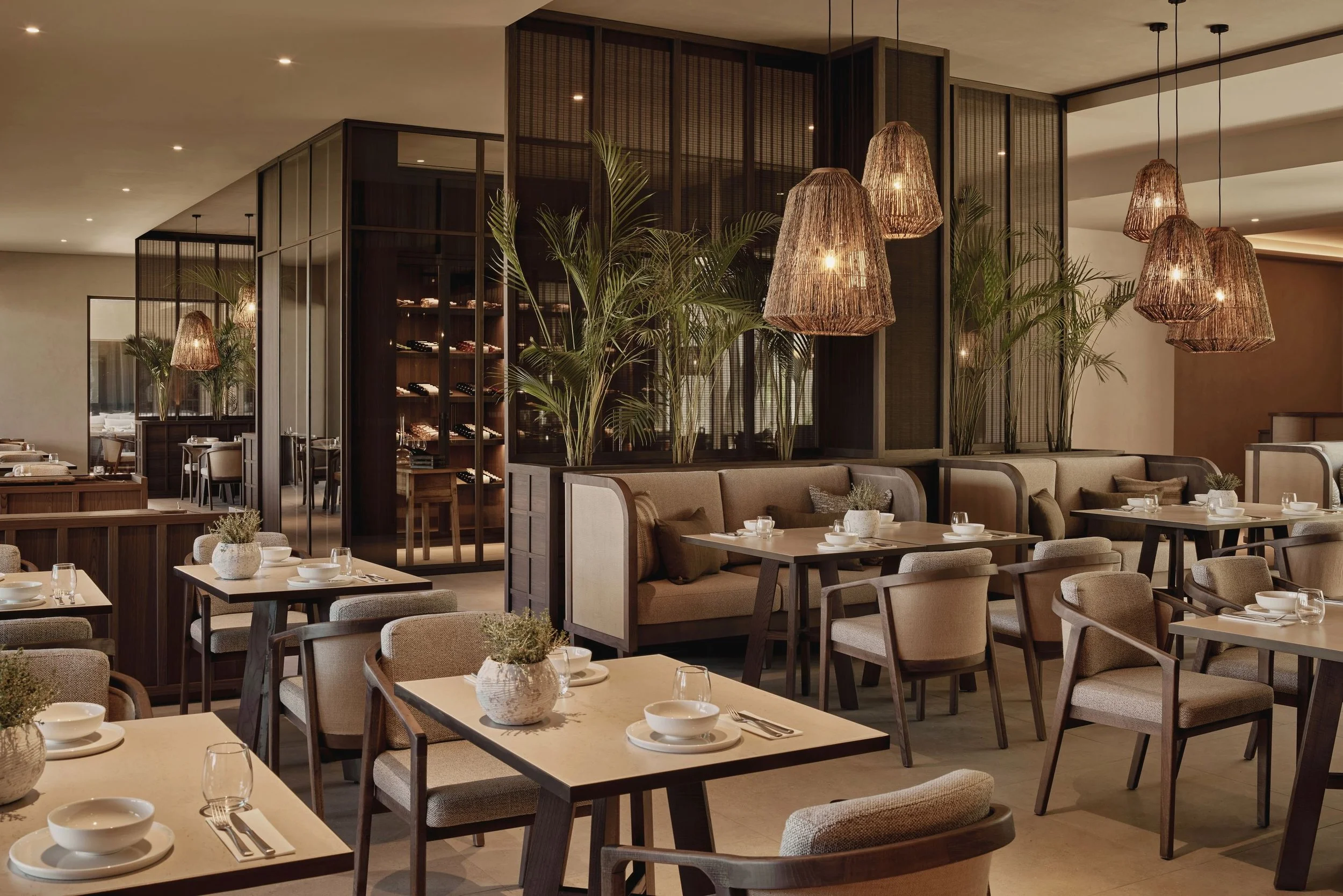 Elegant restaurant interior with beige chairs, wooden tables set with white bowls, glasses, and silverware, large potted plants, and decorative hanging woven light fixtures.