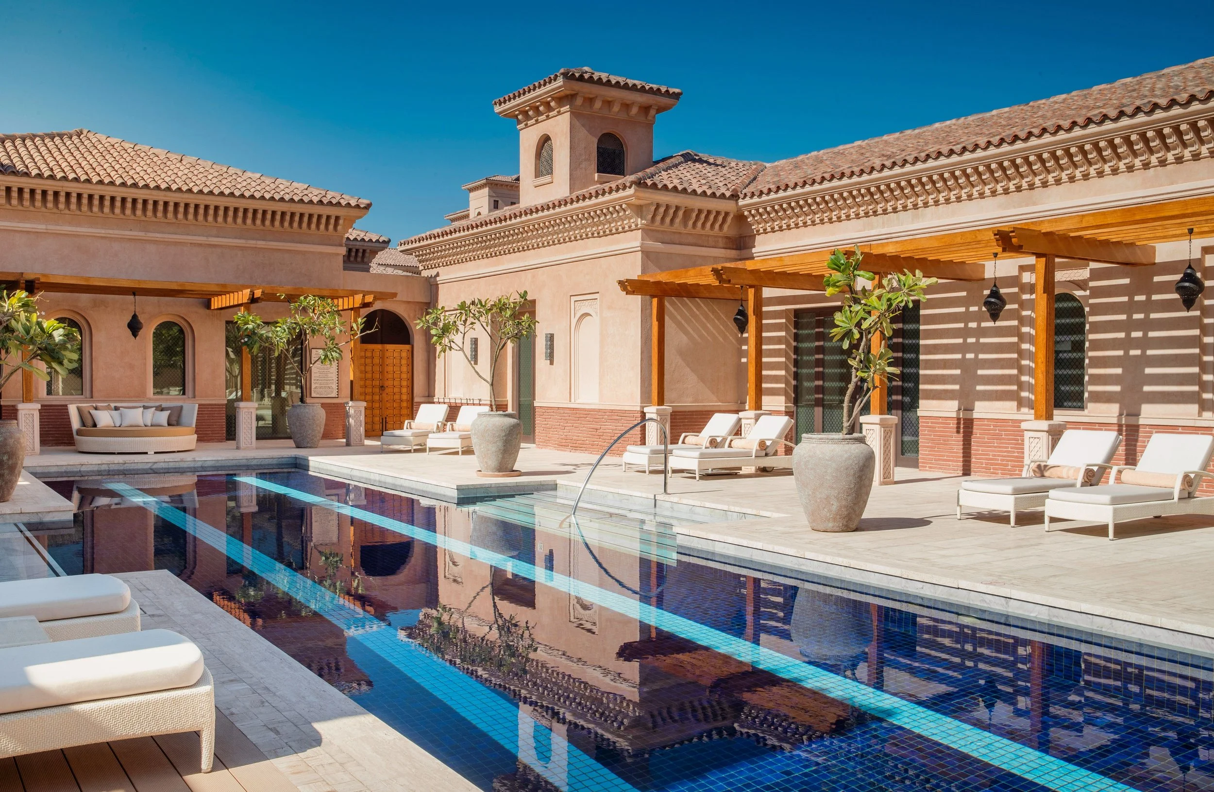 Luxury house with a tiled roof, courtyard, and swimming pool with lounge chairs and potted plants under a clear blue sky.