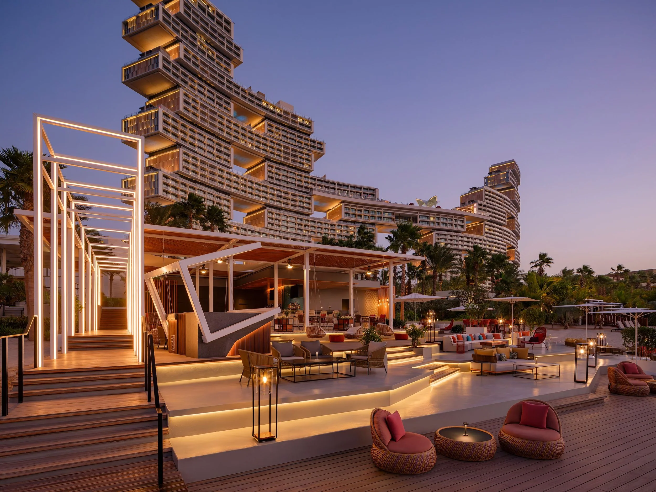 Luxury rooftop bar and lounge area with modern furniture, illuminated stairs, palm trees, and a tall, uniquely designed building in the background during dusk.