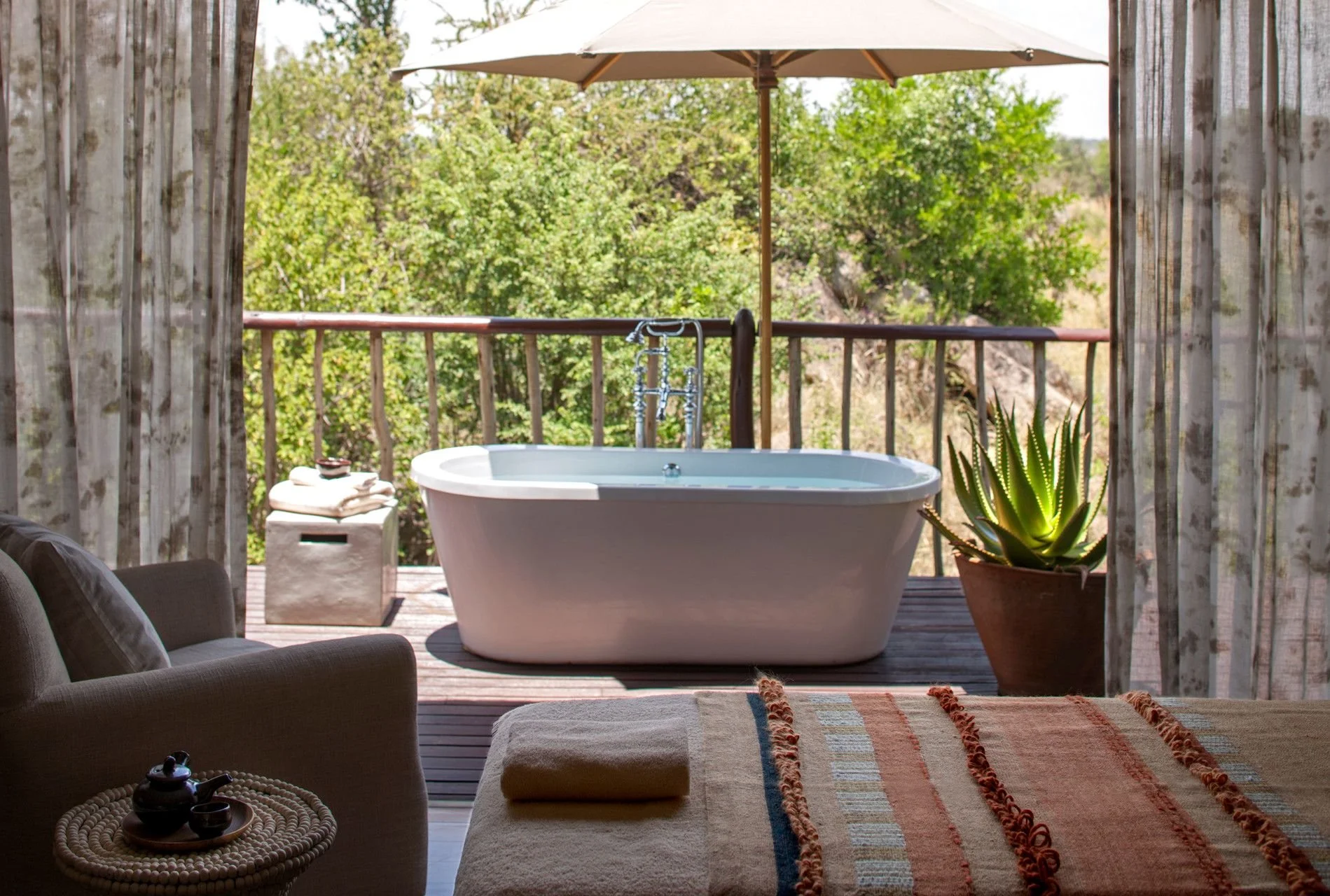 Outdoor balcony with a bathtub, large umbrella, potted plant, and surrounding trees.