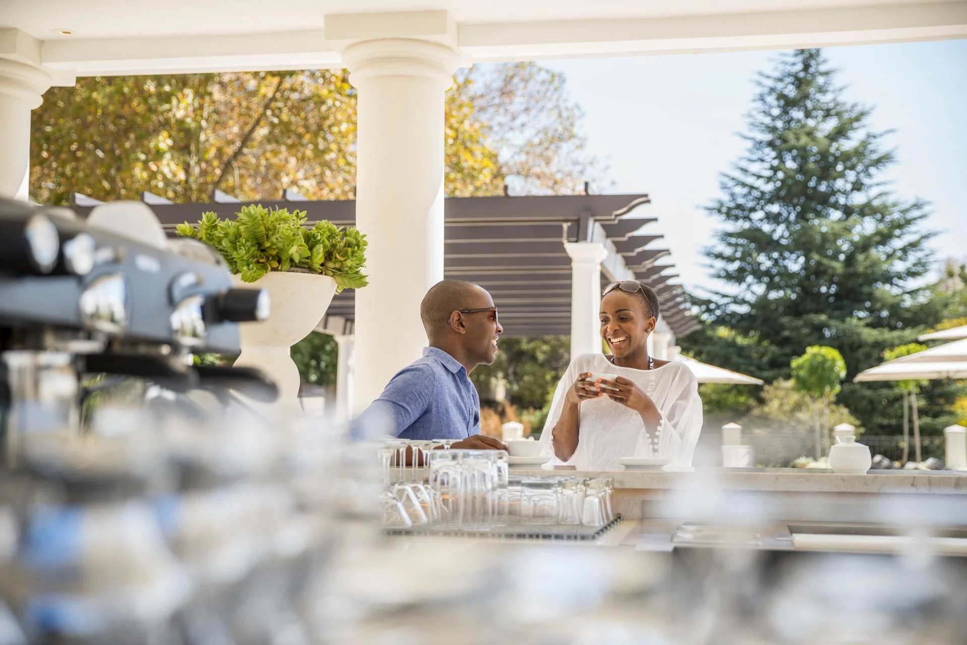 A man and woman enjoying a conversation at an outdoor restaurant table, with a buffet setup in the foreground and trees in the background.