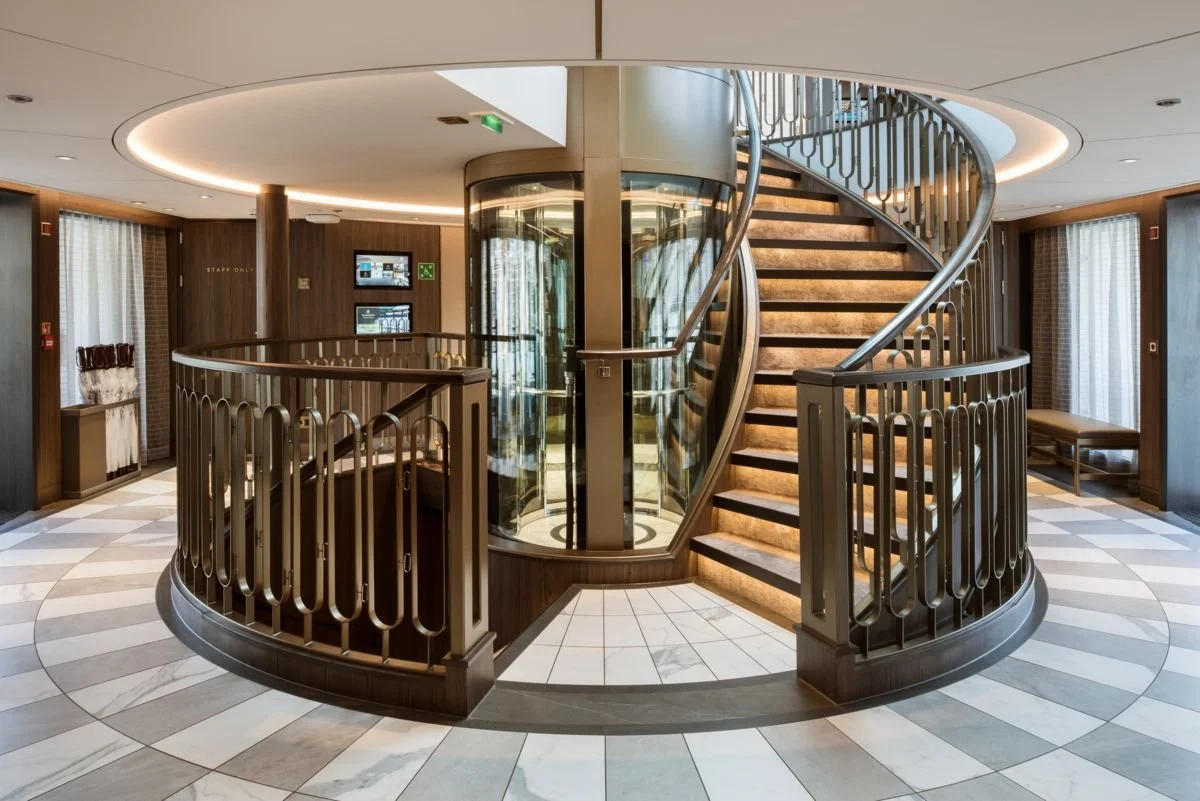 Interior of a modern building with a circular staircase, glass elevator, and patterned tile floor.