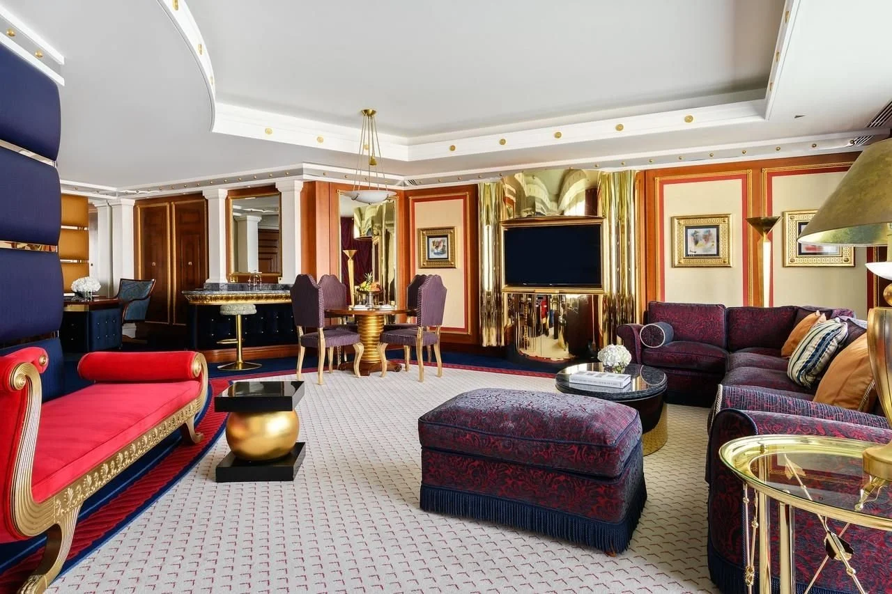 Luxurious hotel living room with patterned carpet, velvet sofas, gold accents, and a flat-screen TV mounted on a gold-paneled wall.