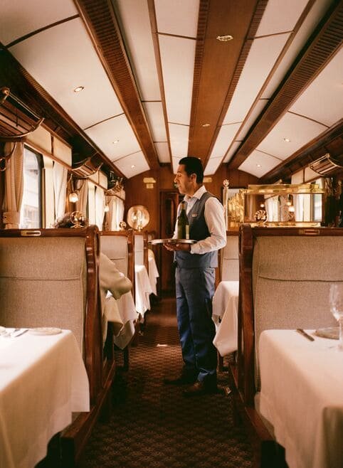 A waiter standing in the aisle of a vintage train dining car, holding a tray with dishes, surrounded by tables with white tablecloths and beige upholstered booths.