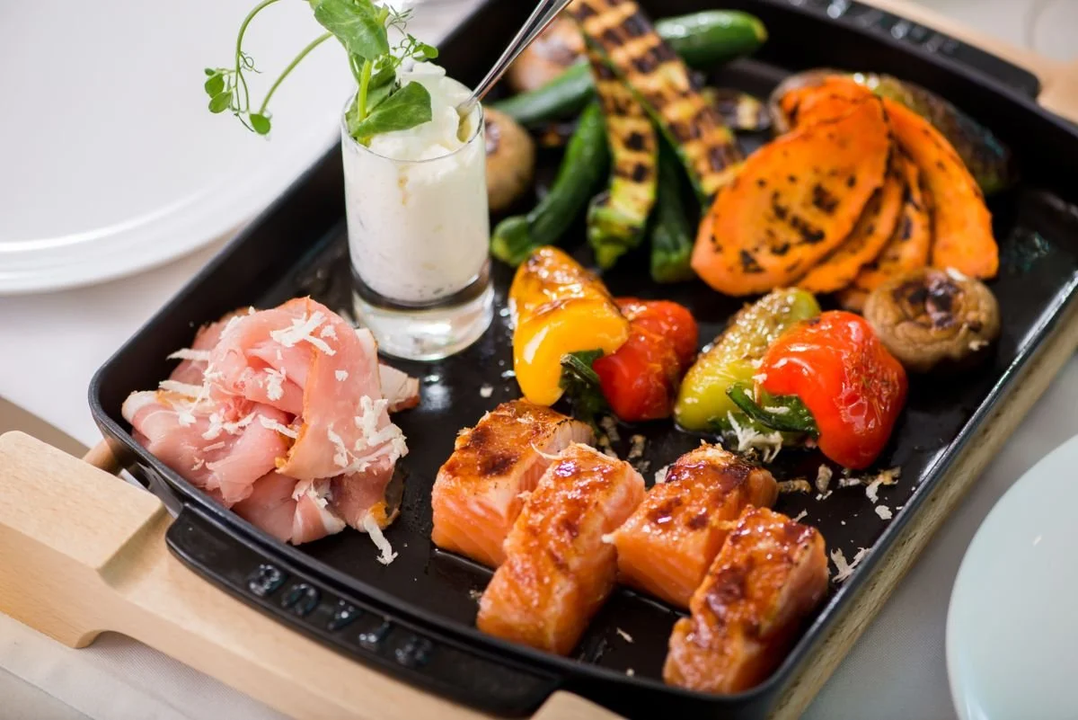 Assorted grilled vegetables, salmon, and prosciutto on a black grill pan with a small cup of dipping sauce and garnished with herbs.