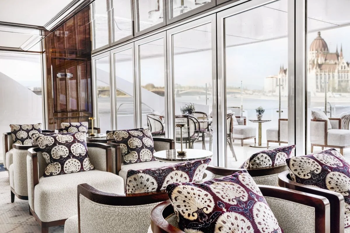 A luxurious indoor lounge with chairs and pillows near large glass windows, overlooking a river and a distant castle.