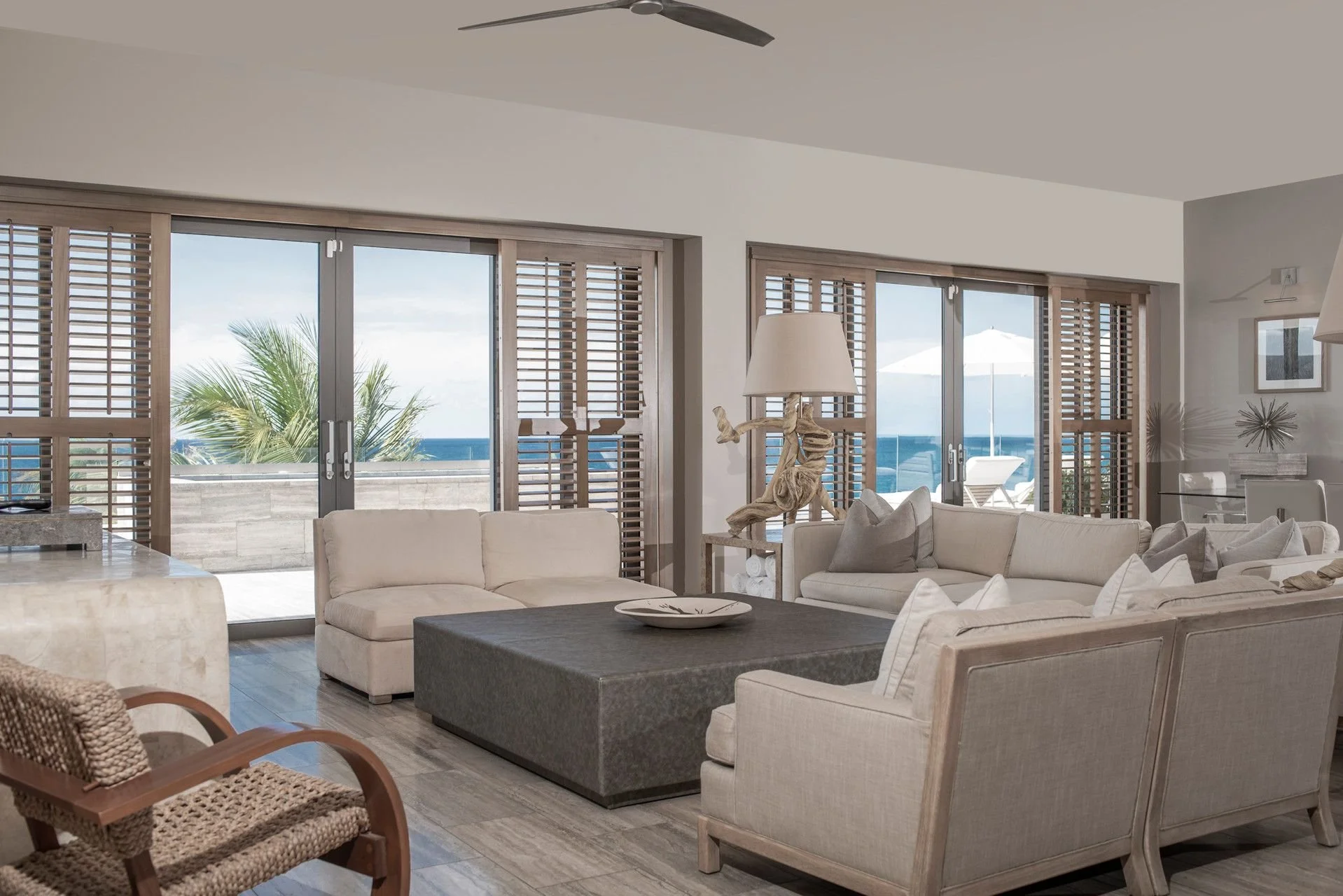 Living room with large glass sliding doors revealing a balcony with ocean view, palm trees, and outdoor umbrella, decorated with neutral-toned furniture, wooden blinds, and tropical decor accents.