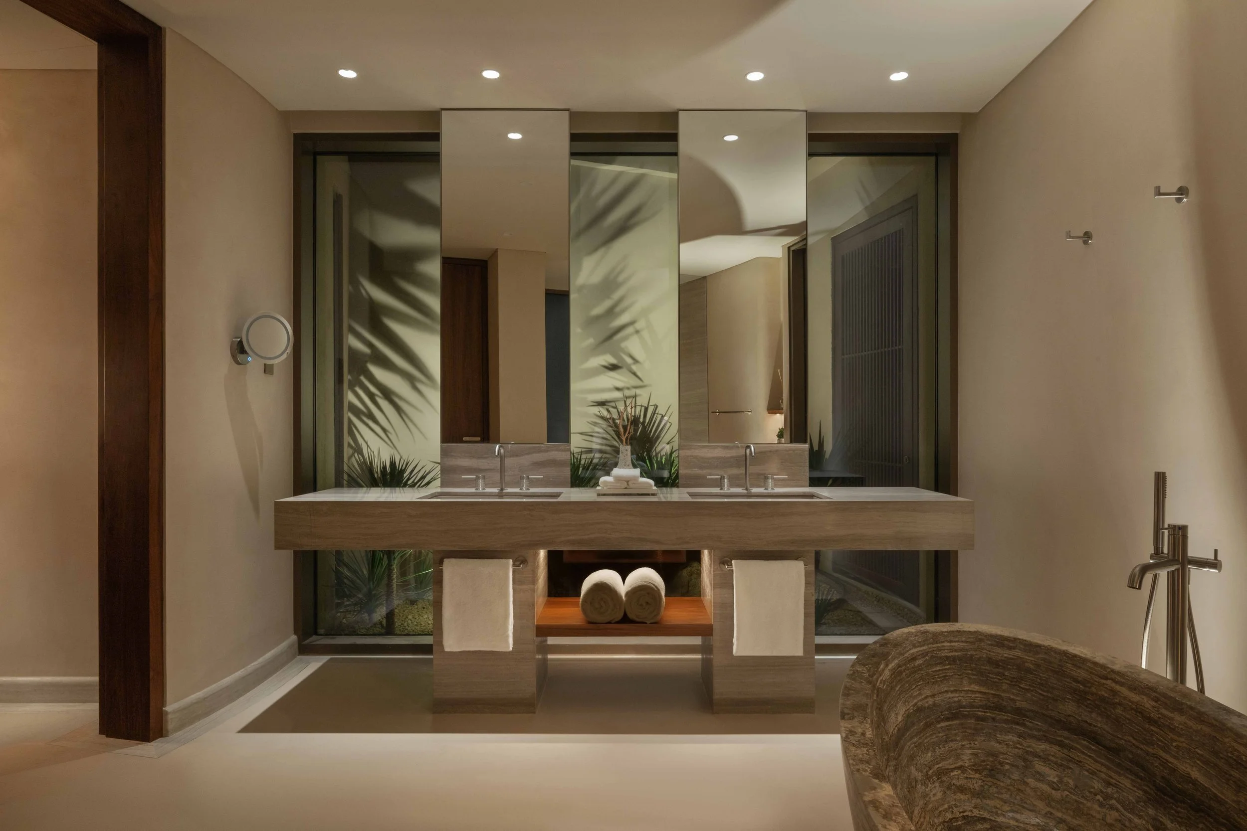 Modern bathroom with double sink vanity, large mirror, beige walls, ceiling lights, and decorative plants.