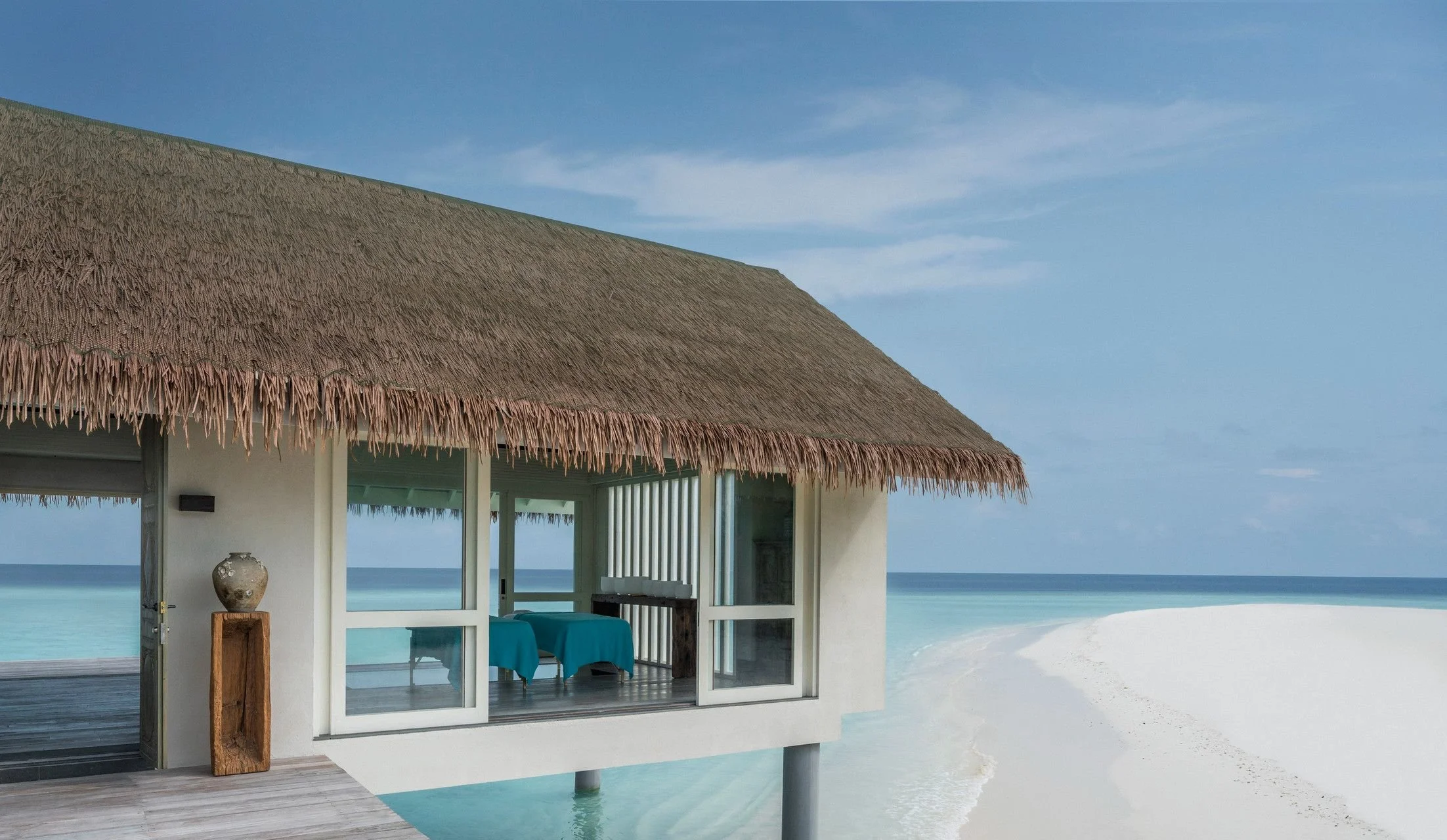 Beachside hut with thatched roof overlooking the ocean, with an open patio area featuring a table with a blue cloth and a decorative vase.