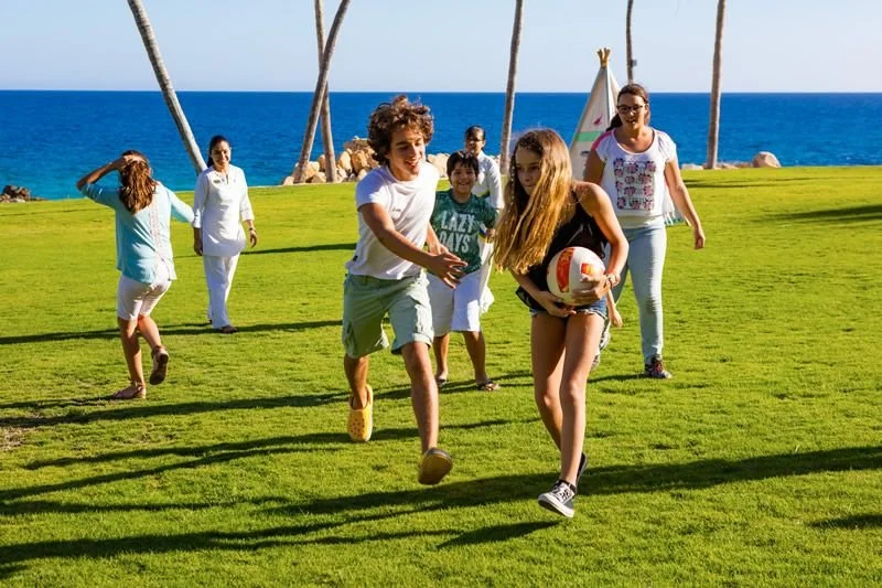 Family playing volleyball on a grassy park near the ocean, with palm trees in the background