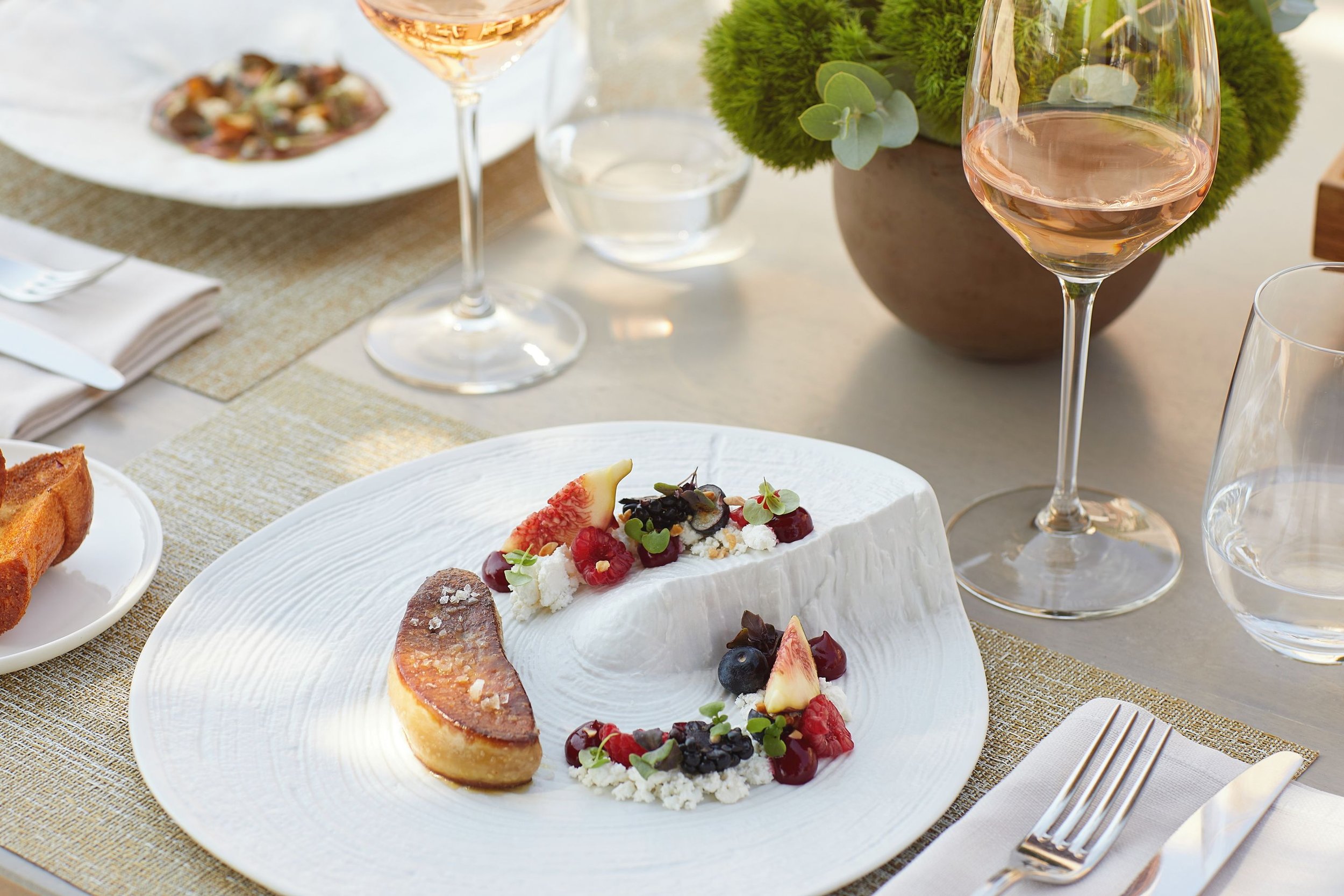 A table set for a meal with white plates, glassware filled with rosé wine, a cheese platter topped with assorted berries and garnishes, and a slice of cake, with a floral centerpiece.