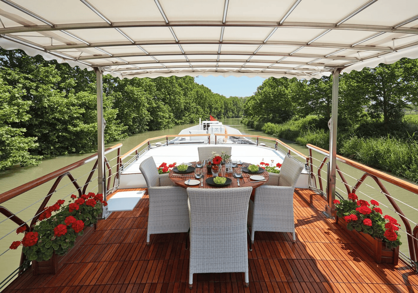A dining setup on the deck of a boat with a round wooden table, six white wicker chairs, and a canopy overhead, overlooking a narrow river surrounded by green trees.
