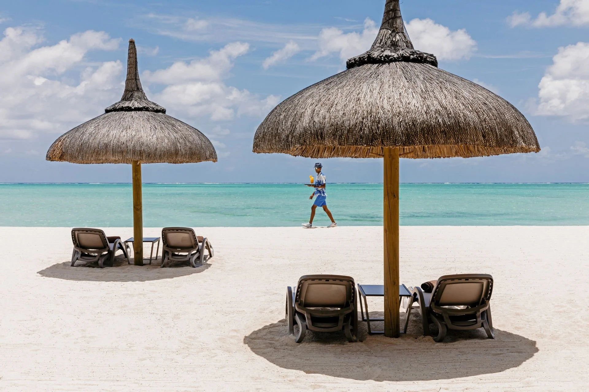 Two thatched beach umbrellas with lounge chairs on a sandy beach overlooking turquoise water, with a man in a light shirt, shorts, and a hat walking along the shoreline under a partly cloudy sky.