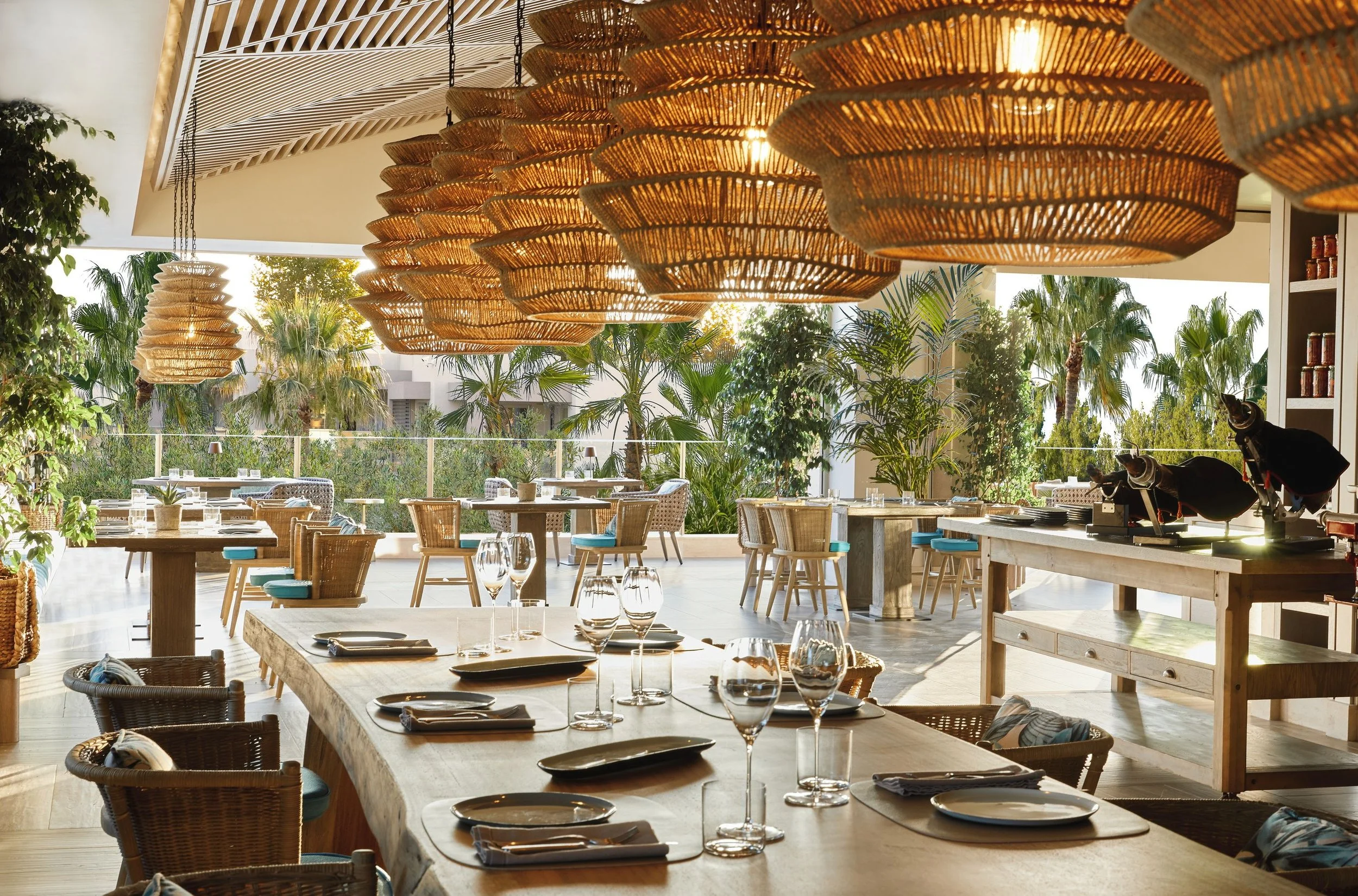 Indoor restaurant with wooden tables set with glassware, plates, and napkins, decorated with hanging wicker light fixtures and surrounded by lush green plants and palm trees outside.