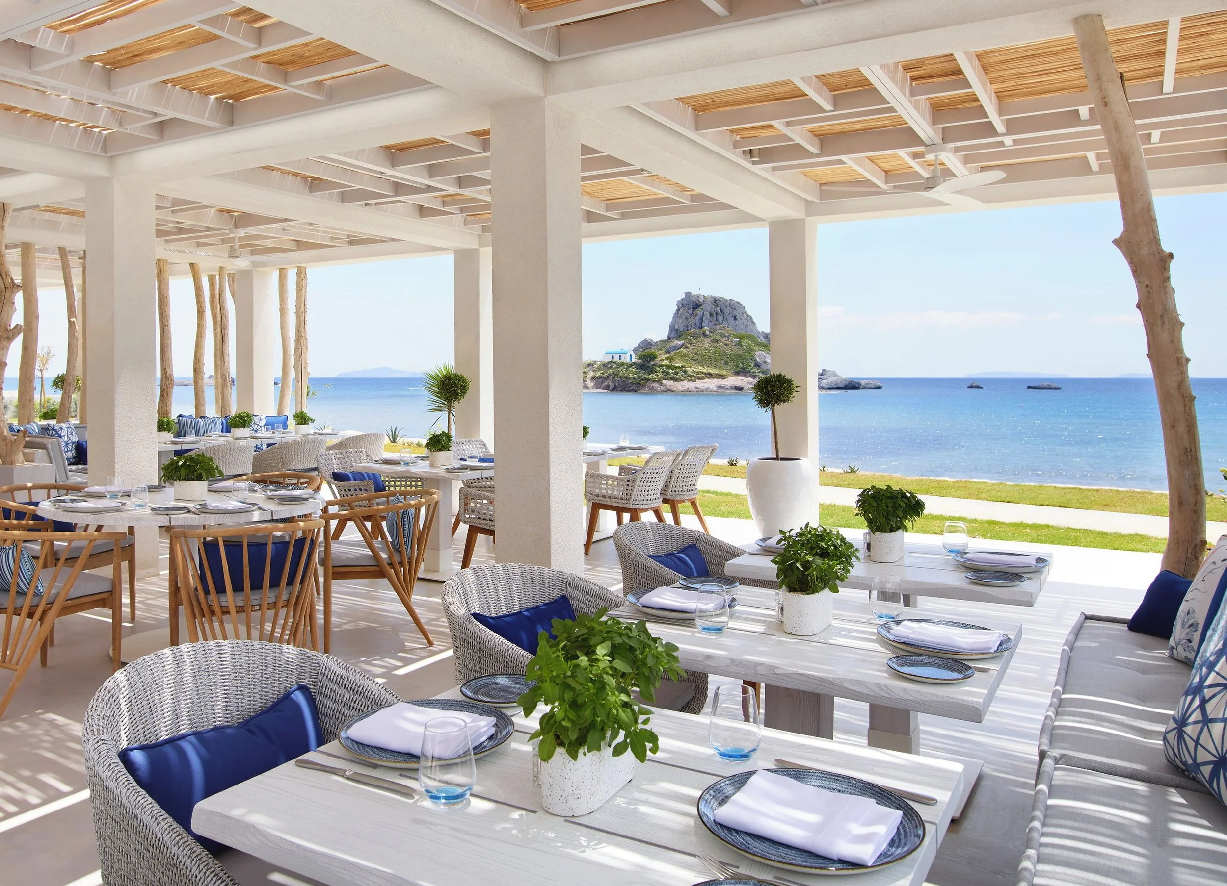 Outdoor dining area with white tables and chairs, potted plants, and ocean view with distant island and rock formations.