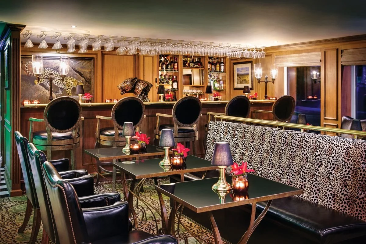 A sophisticated bar area with dark wood paneling, black leather chairs, leopard print accents, and a variety of liquor bottles on shelves. Glasses hang upside down from above, and small tables with lamps and pink flowers are arranged for seating.
