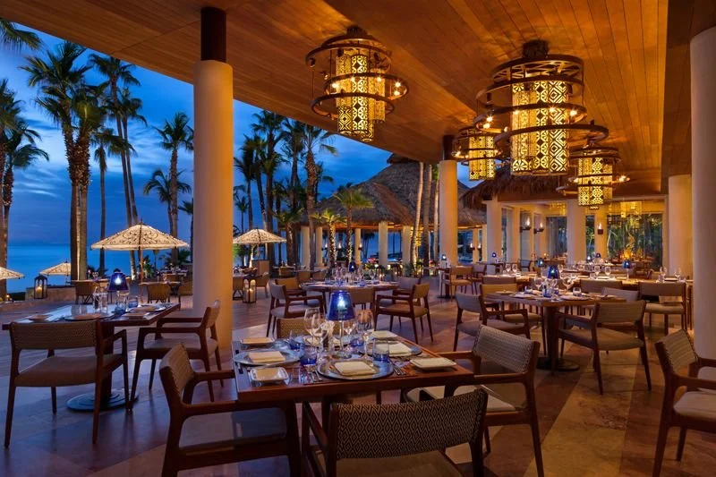 Elegant outdoor seaside restaurant dining area at dusk with palm trees, thatched umbrellas, and hanging lantern-style lights.