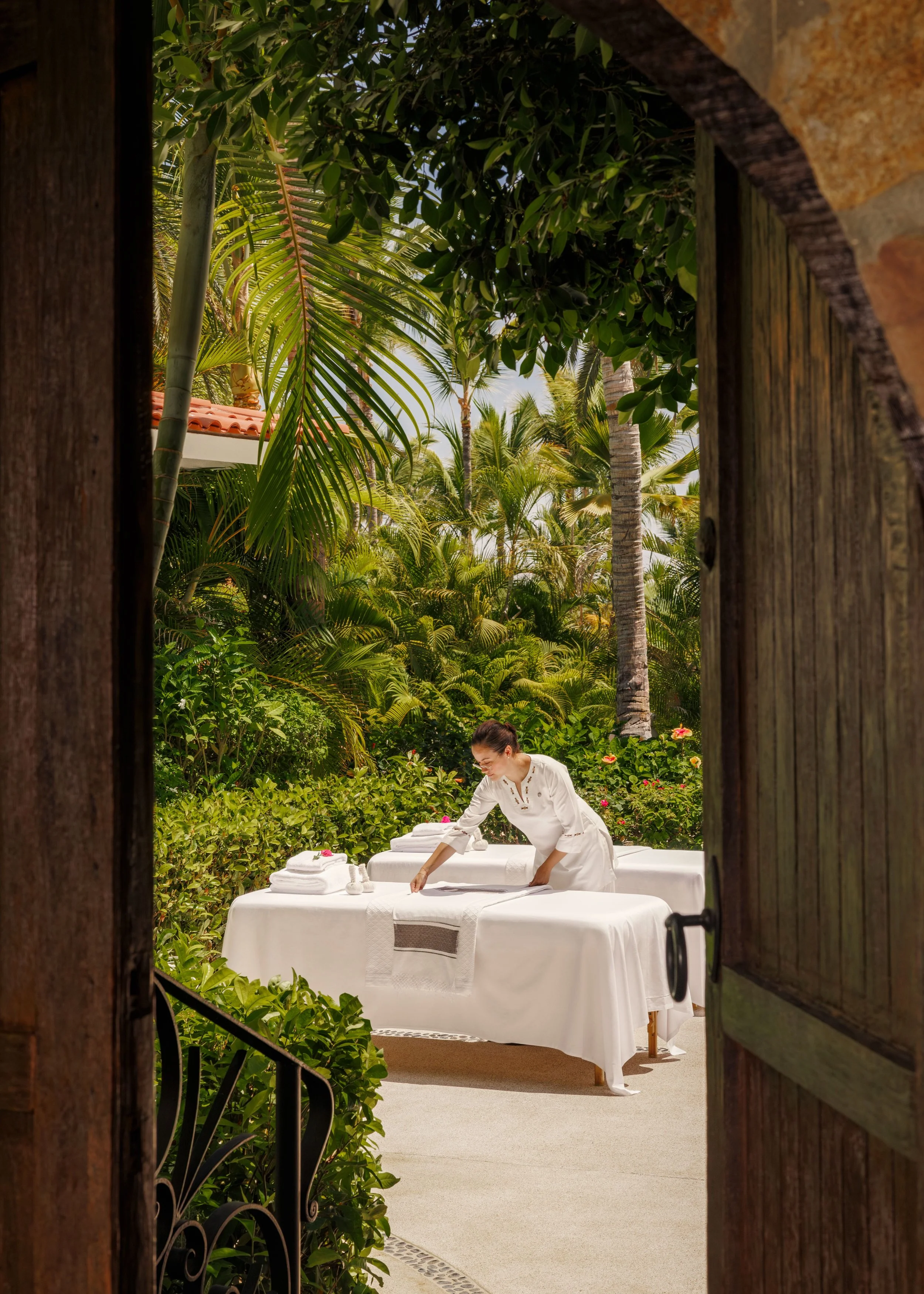 View through an open wooden gate showing a woman preparing bedsheets on a massage table outdoors in a lush garden with tall palm trees and tropical plants.