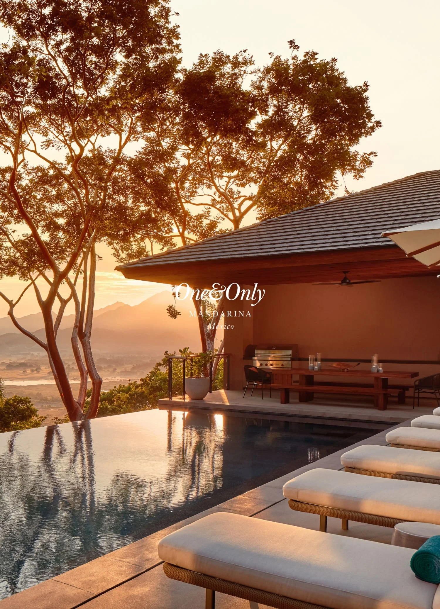 A luxurious outdoor scene during sunset featuring a rooftop infinity pool, white lounge chairs with blankets, a shaded patio with a wooden table and chairs, a grill, and tall trees with orange leaves against a scenic view of mountains.