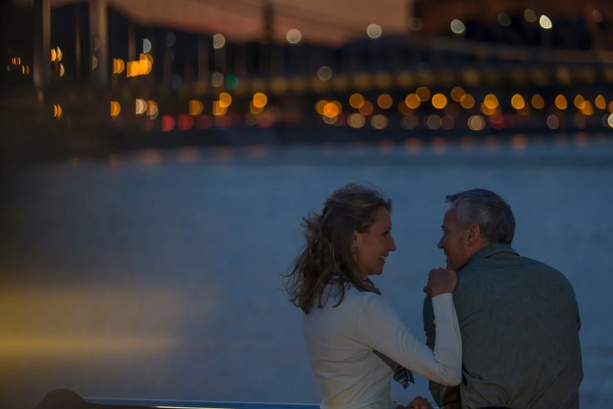 A woman and a man are sitting closely by a body of water at dusk, smiling and looking at each other, with city lights blurred in the background.
