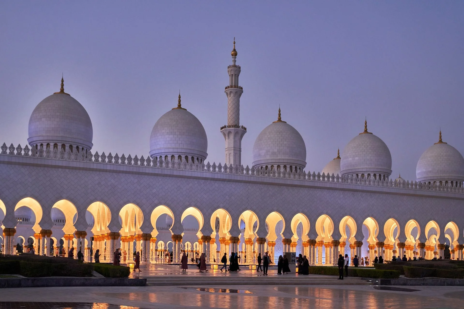 The Sheikh Zayed Grand Mosque illuminated at dusk, showing white domes, minarets, and arched colonnade with visitors walking beneath.