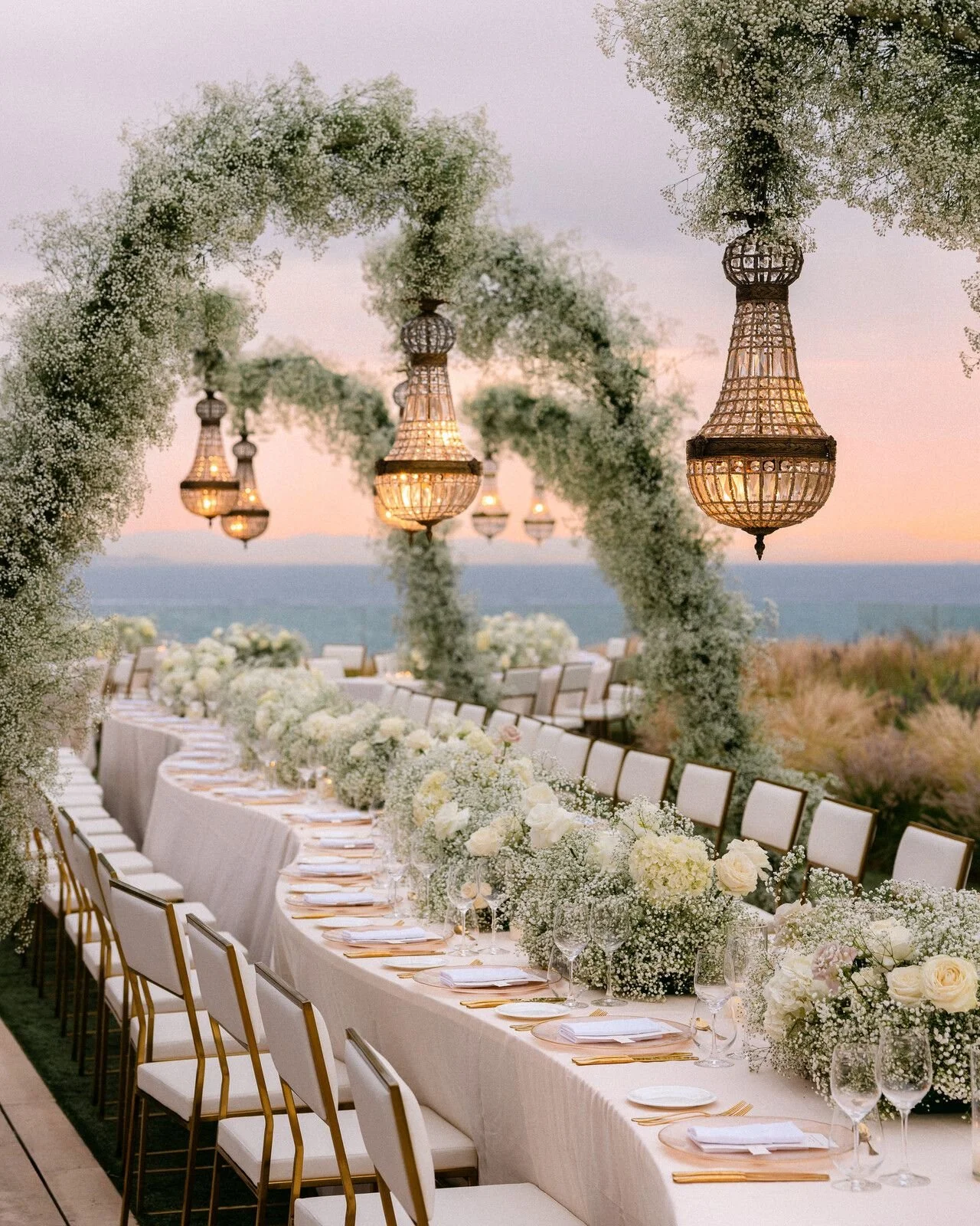 Elegant outdoor wedding reception table with white floral arrangements and hanging chandeliers at sunset.