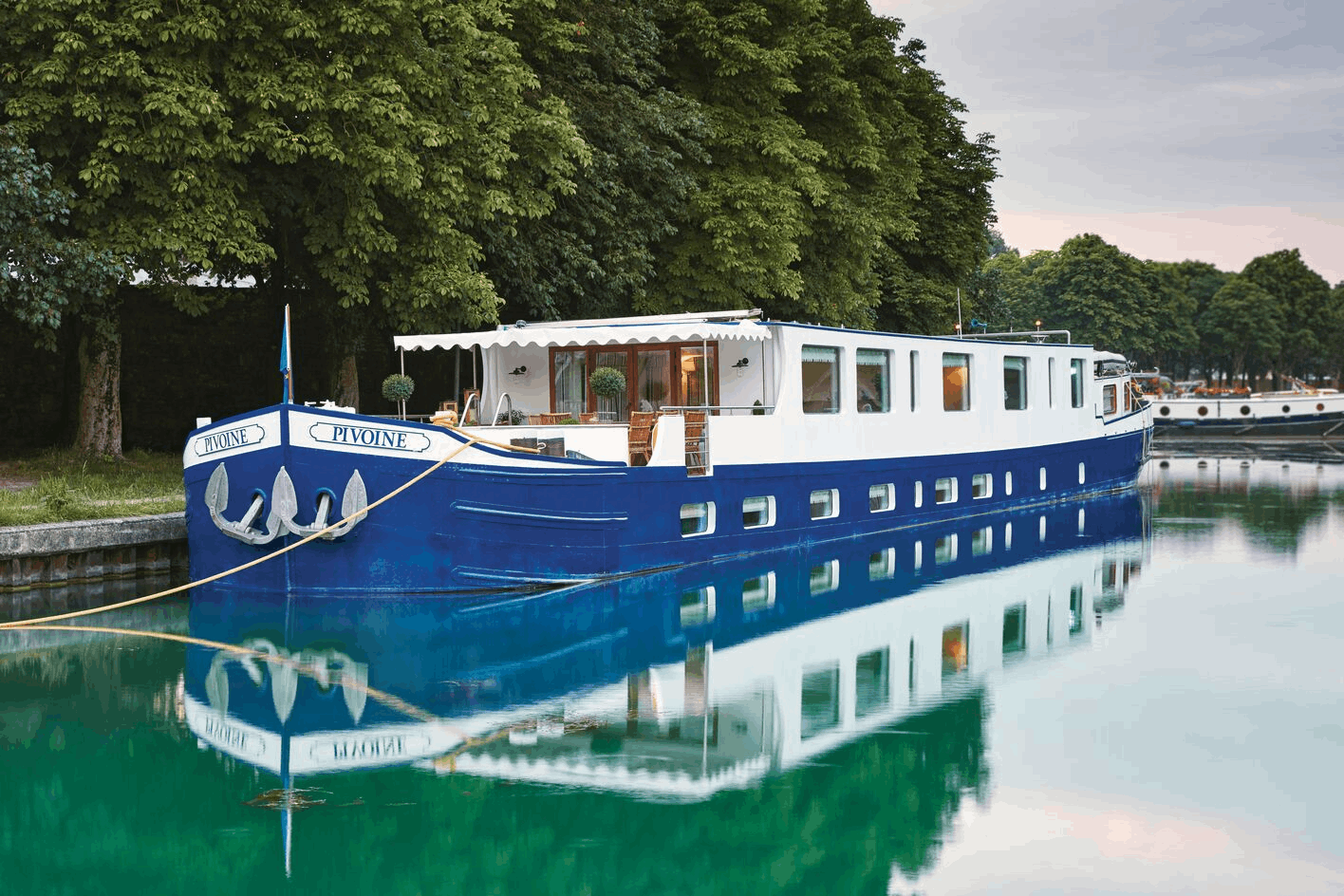 A blue and white houseboat named Pivoine docked on calm water with green trees in the background.