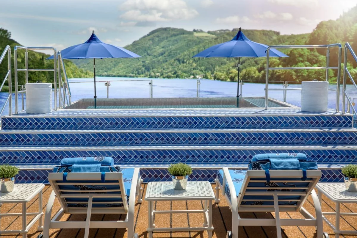 Swimming pool with blue tiled steps leading to the water, surrounded by lounge chairs, small tables, and potted plants, overlooking a river and green hills under a partly cloudy sky.
