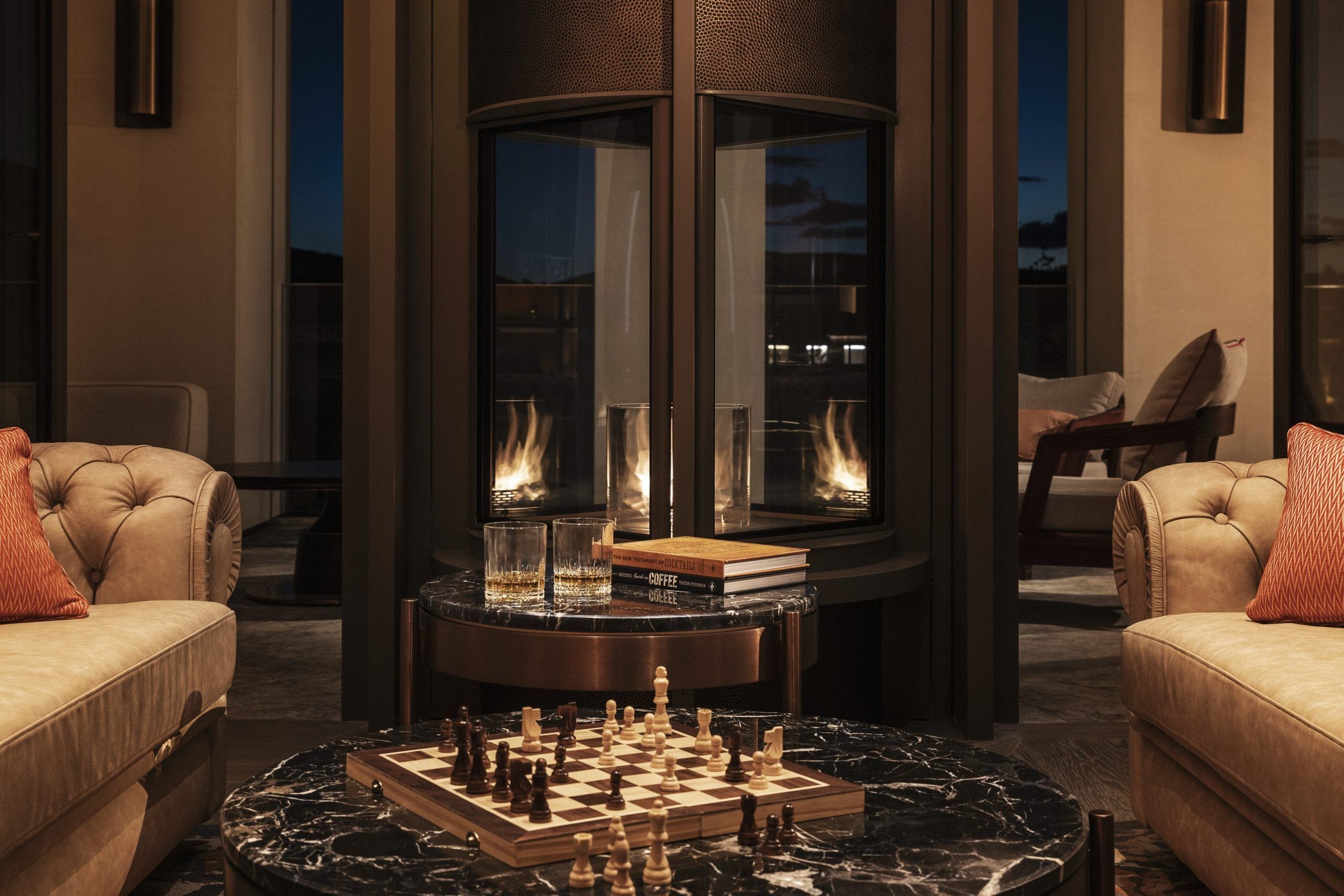 A cozy living room with beige sofas, a black marble coffee table with a chessboard, and a modern glass fireplace with flames. Behind the fireplace, large windows reveal a nighttime view of distant trees and mountains.