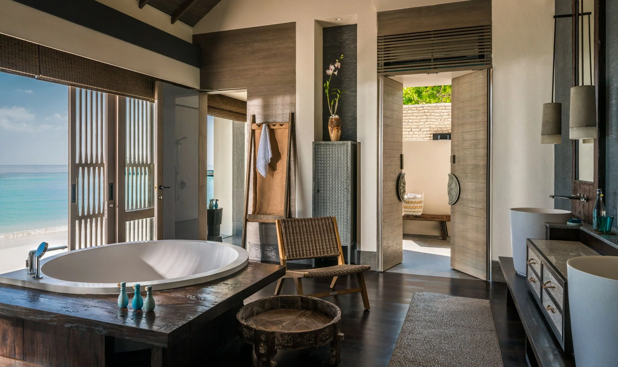 A bathroom with a view of the beach and ocean through sliding glass doors, featuring a modern bathtub, wooden furniture, potted plants, and natural light.
