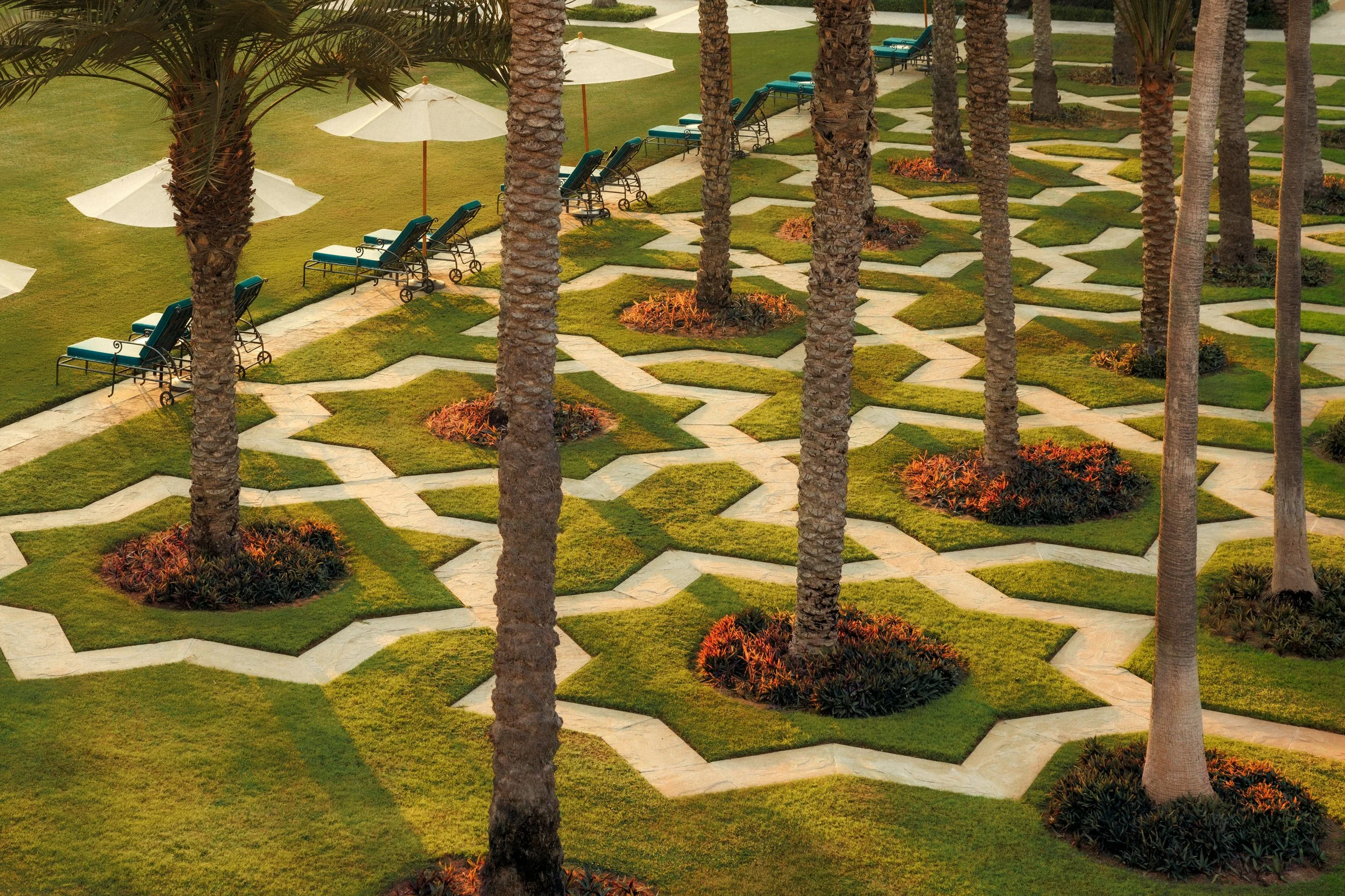 A landscaped outdoor area with palm trees, paved walkways, green grass, and benches with umbrellas.